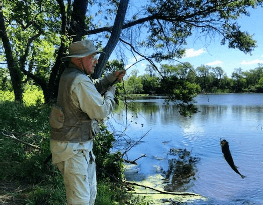 A fisherman in a beige outfit catches a fish by a river under a clear blue sky.