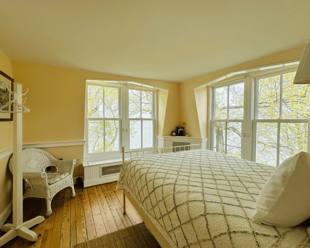 A cozy bedroom with a bed, a wicker chair, and large windows overlooking greenery.