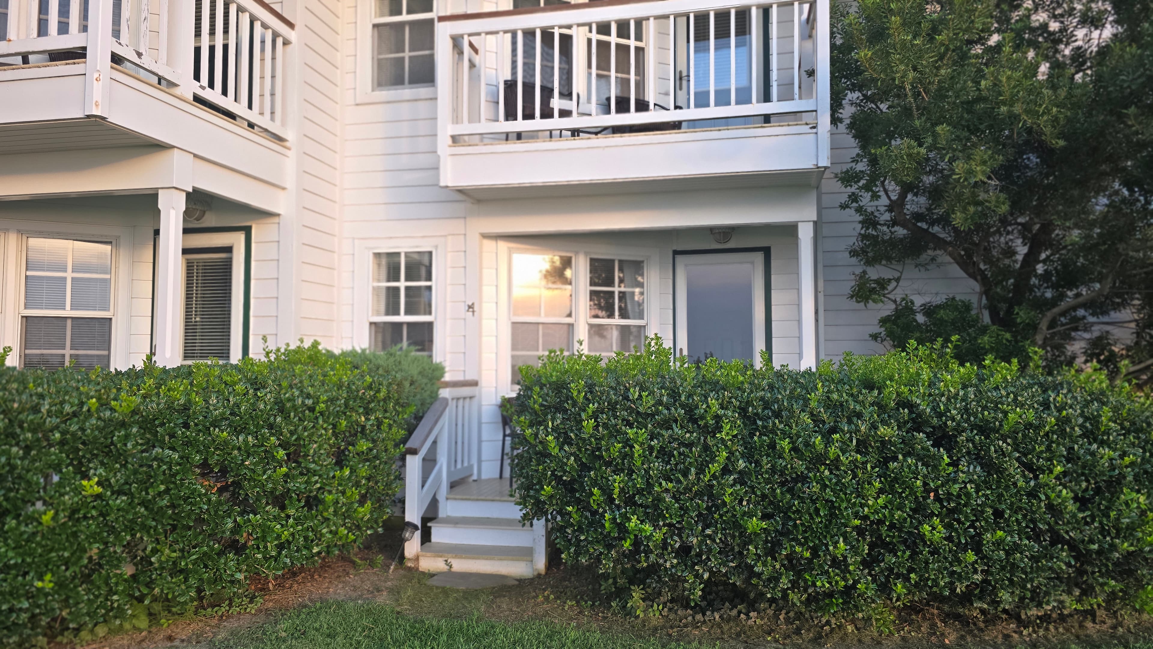 Exterior view of a two-story white house with a staircase and hedge, reflecting sunset light.