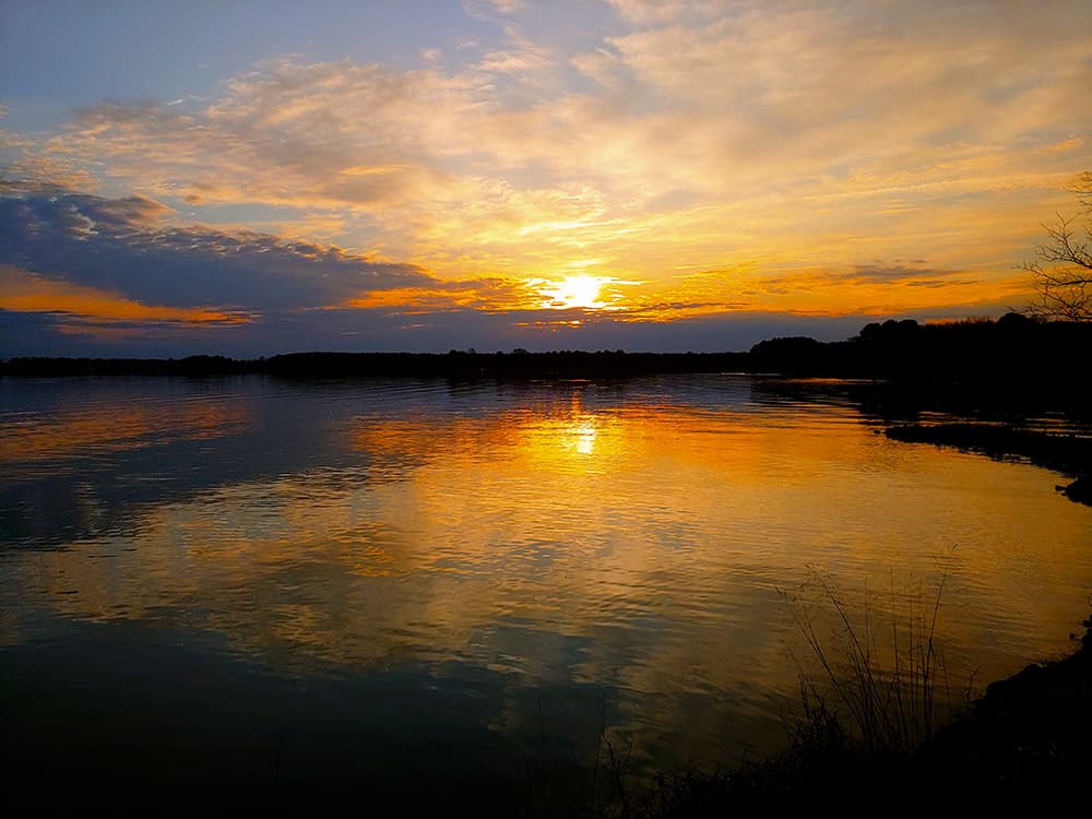 Sunset over a calm lake, reflecting vibrant orange and yellow hues in the water.