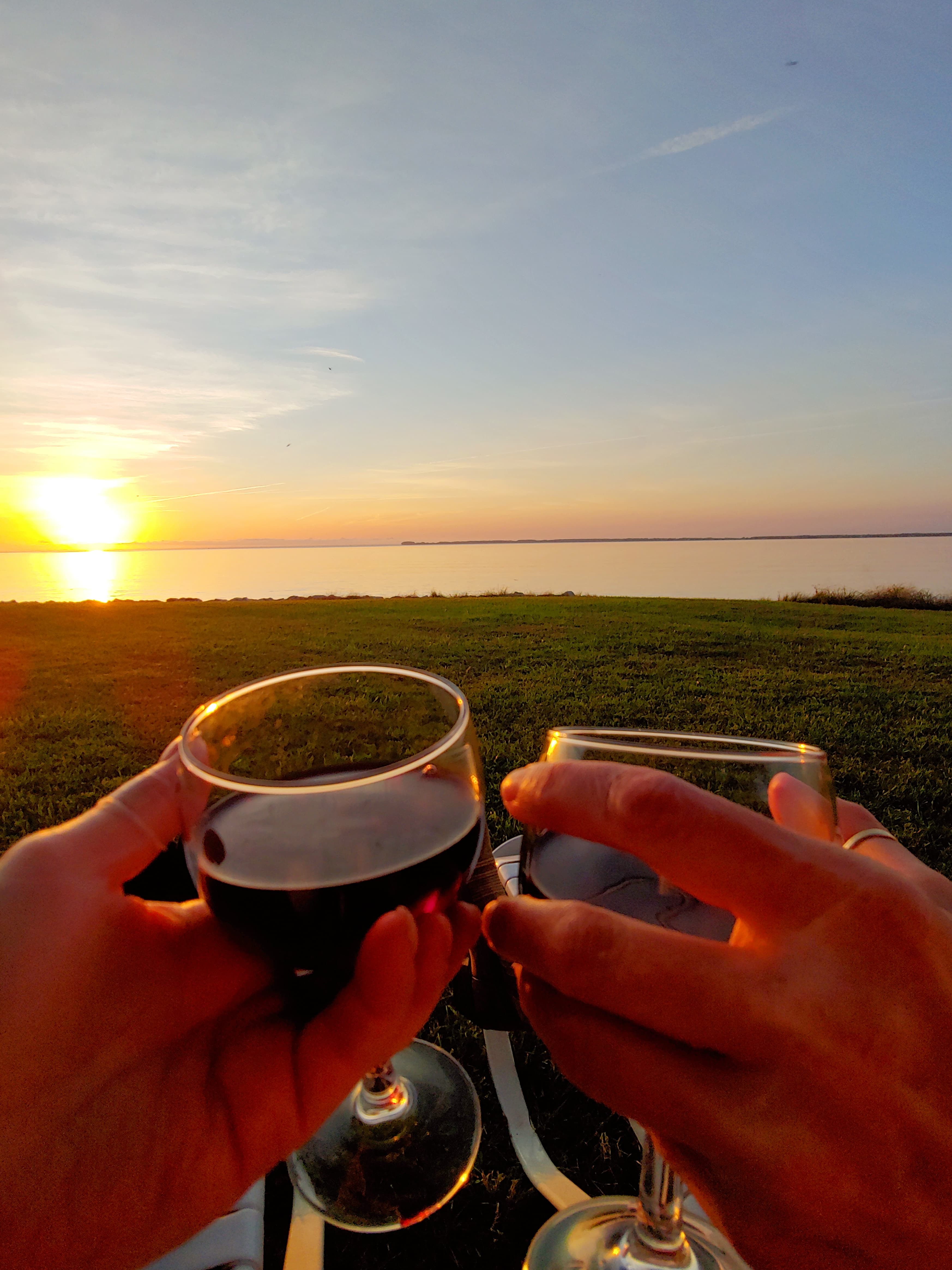 Two hands clink wine glasses against a sunset over calm waters.