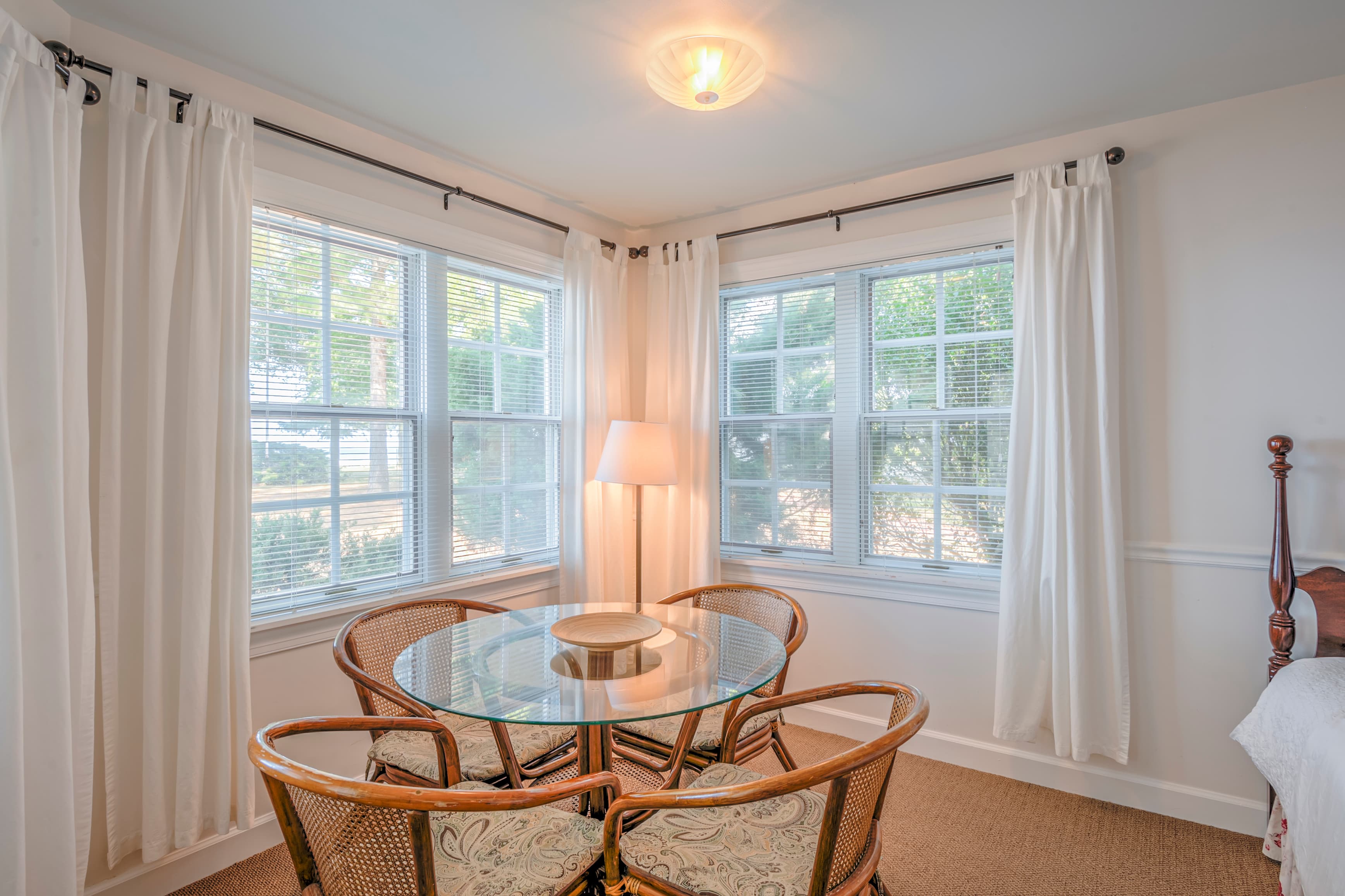 A cozy dining nook with a glass table surrounded by four rattan chairs, illuminated by natural light from large windows.