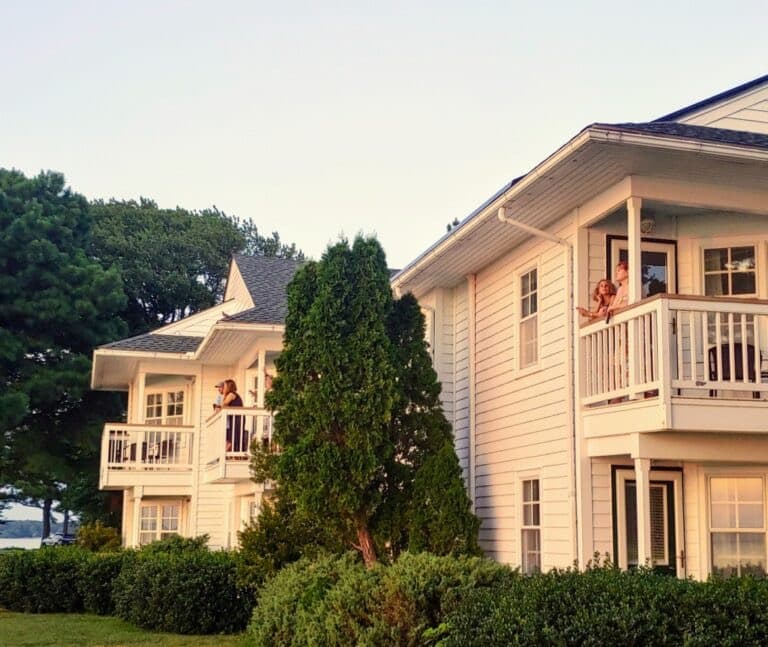 Two people are seen on balconies of white houses, surrounded by green trees and bushes.