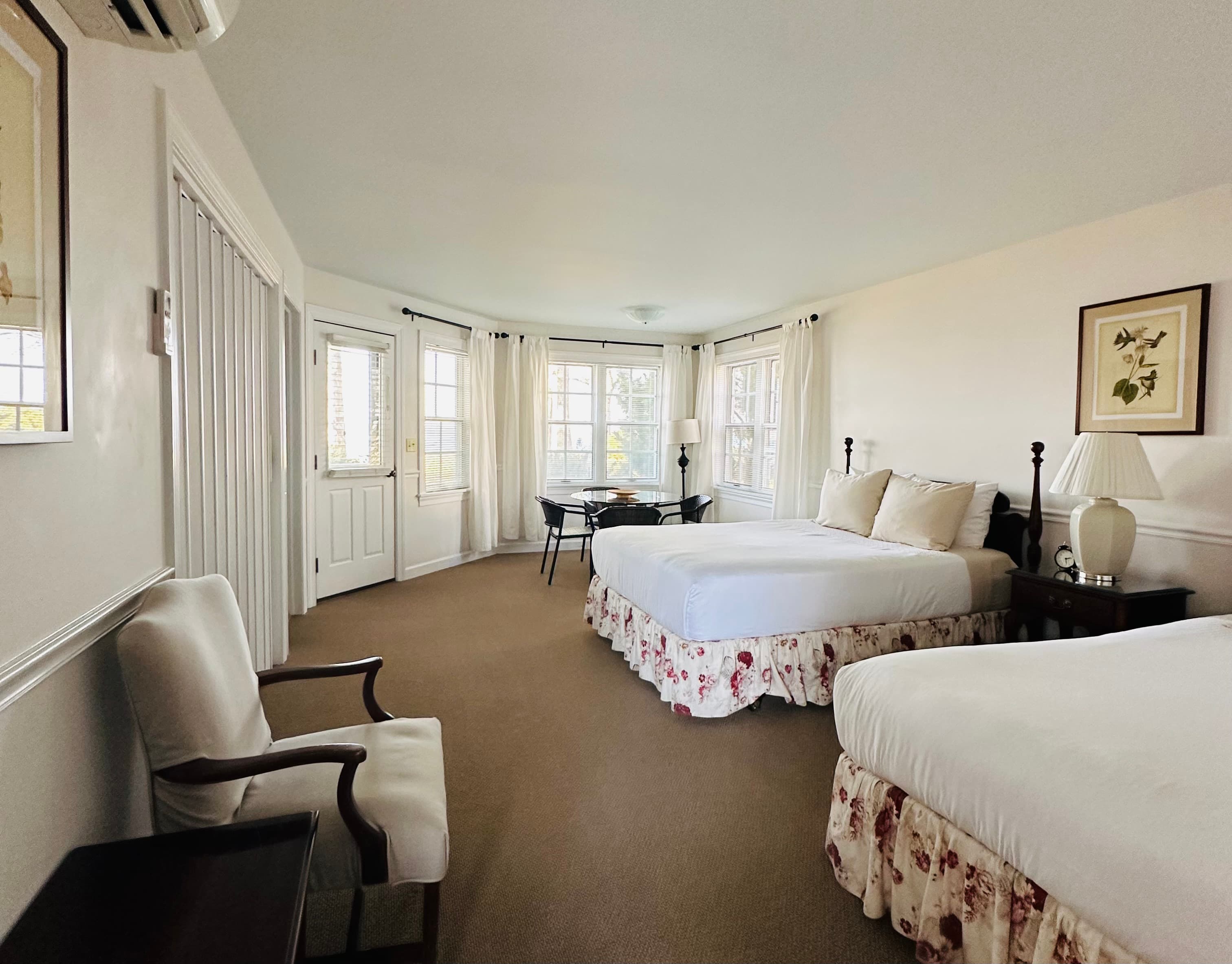 A bright hotel room featuring two beds, a table, and large windows with curtains.