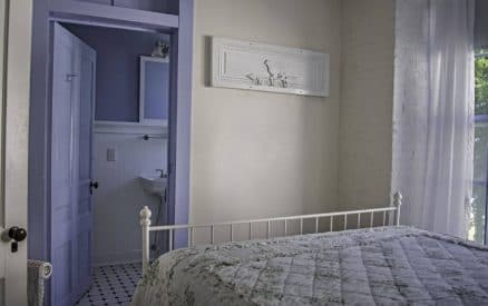 A cozy bedroom featuring a white metal bed and a view into a small purple-walled bathroom.