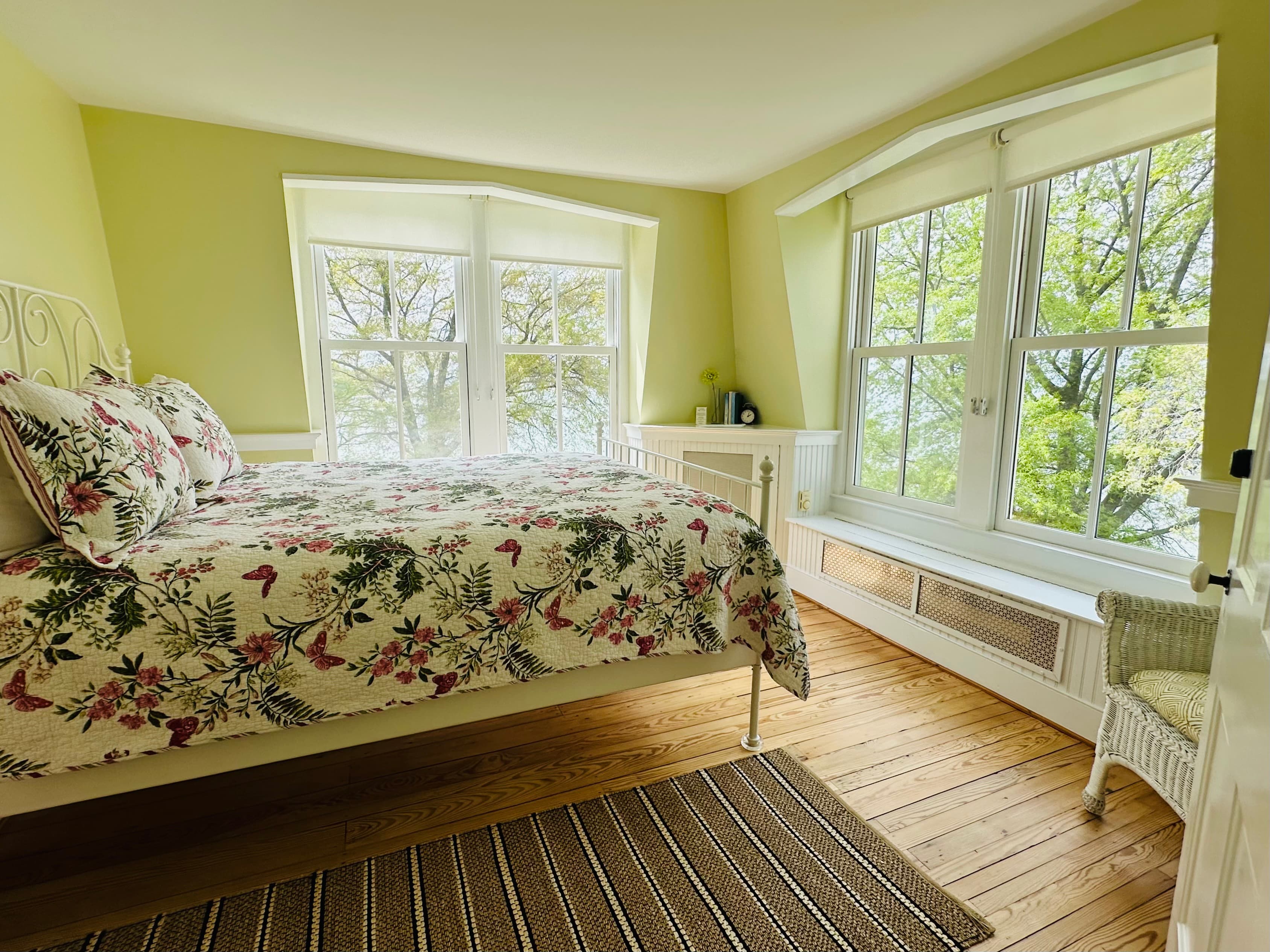 A bright bedroom features a floral-patterned bedspread, large windows, and a cozy reading nook.