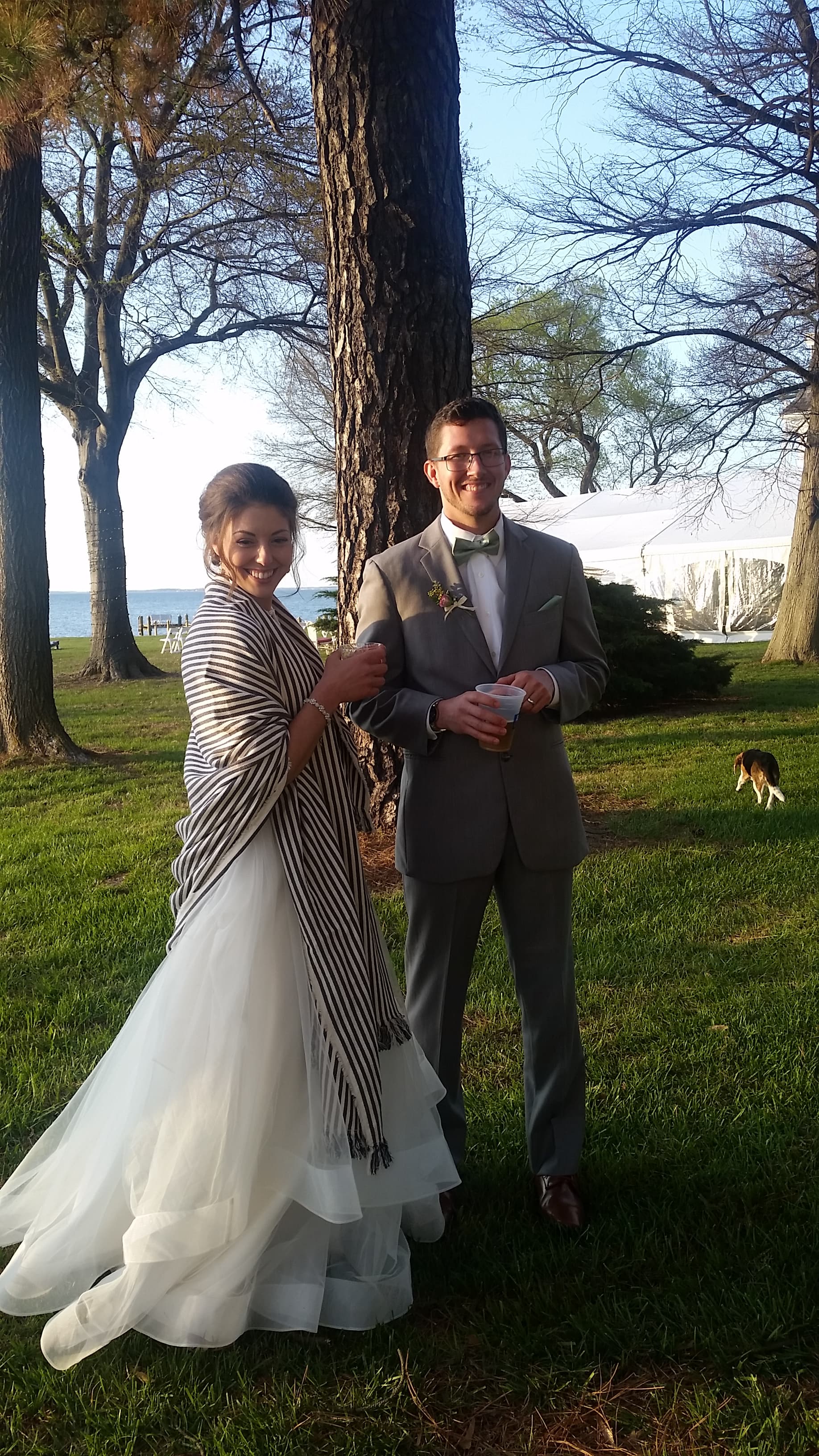 A bride and groom stand together outdoors, smiling, with trees and a lake in the background.
