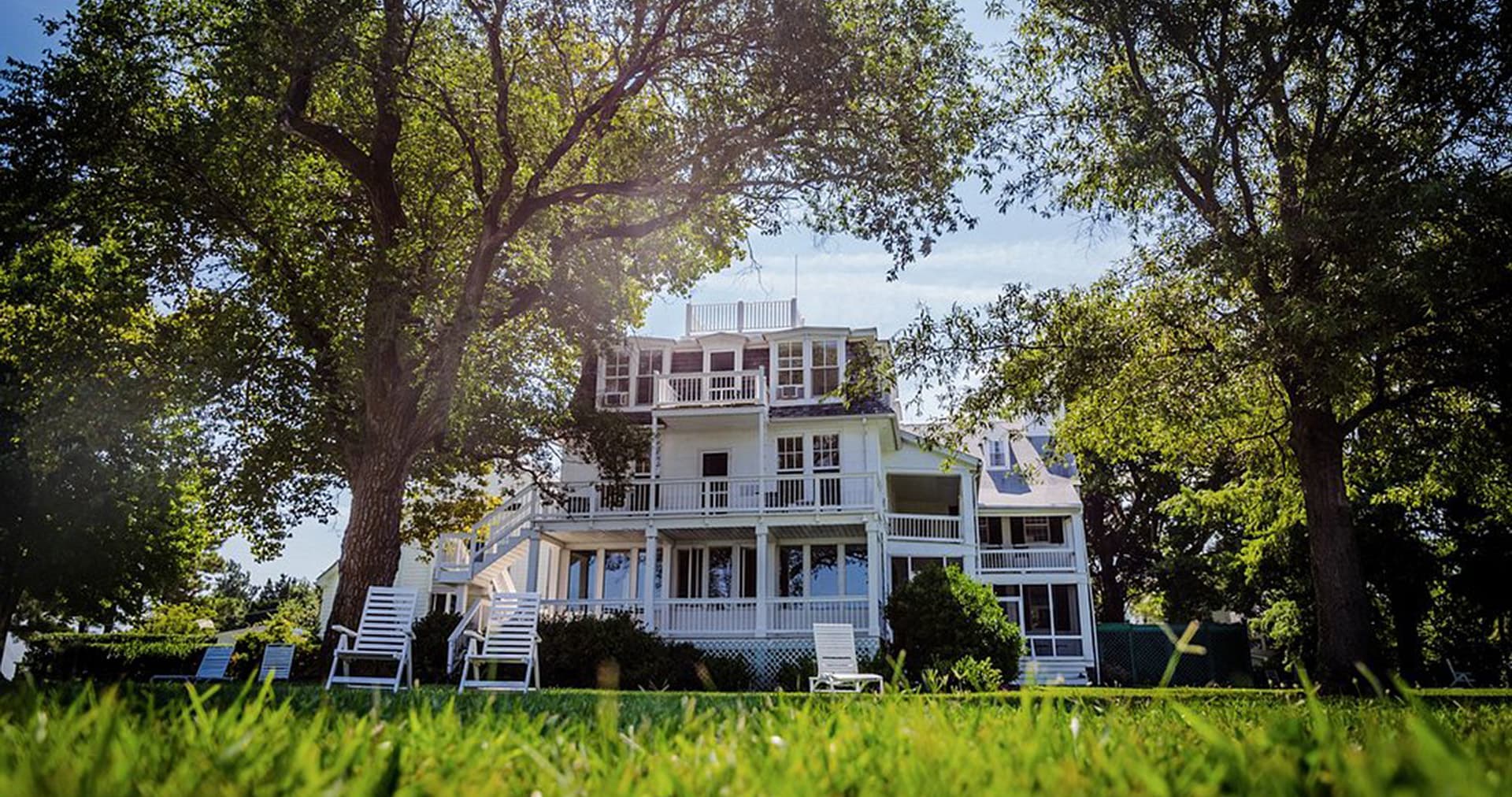 A large white house surrounded by trees and lawn chairs on a sunny day.