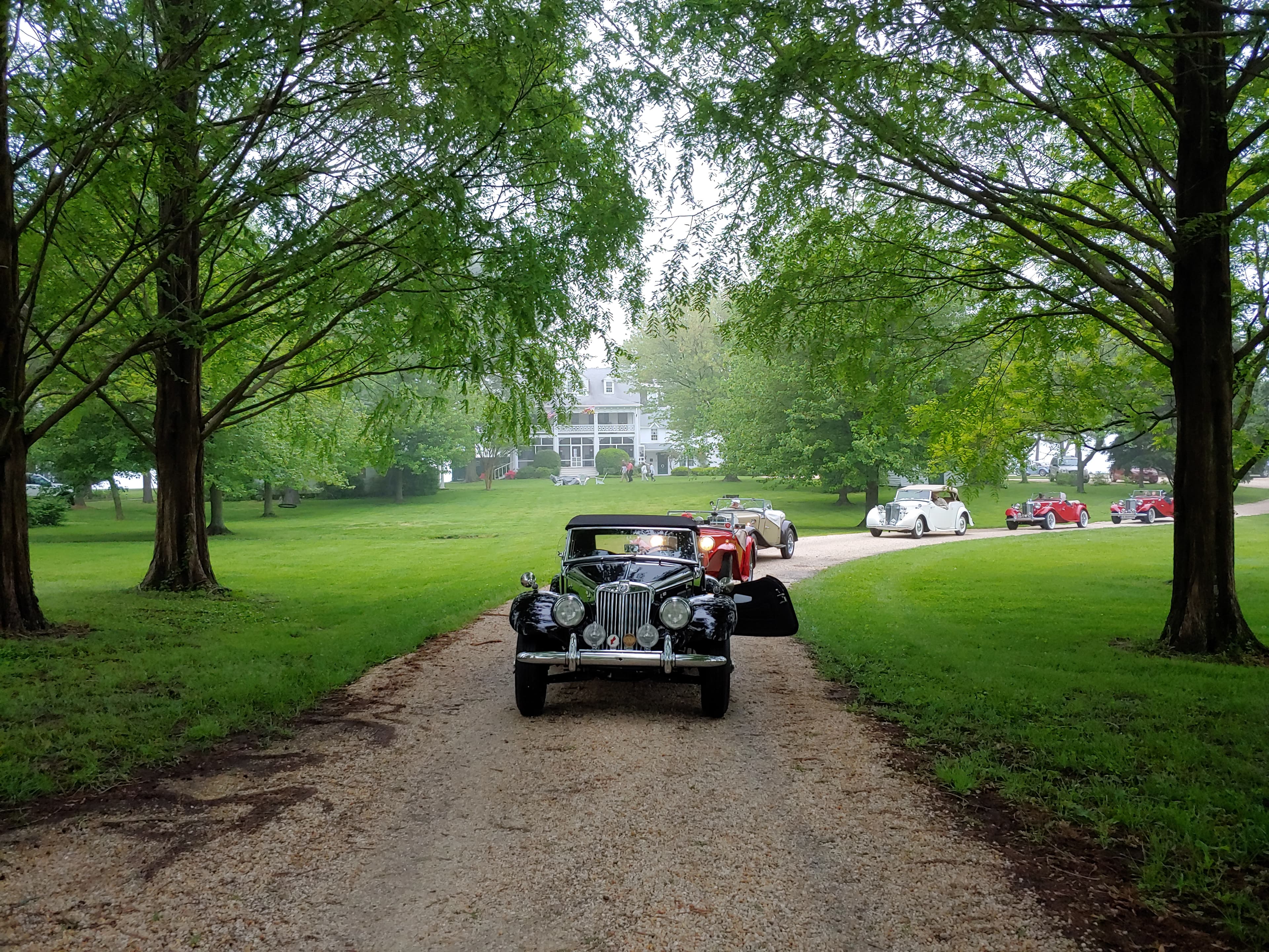 A vintage car parked on a gravel driveway, surrounded by lush greenery and an elegant house in the background.