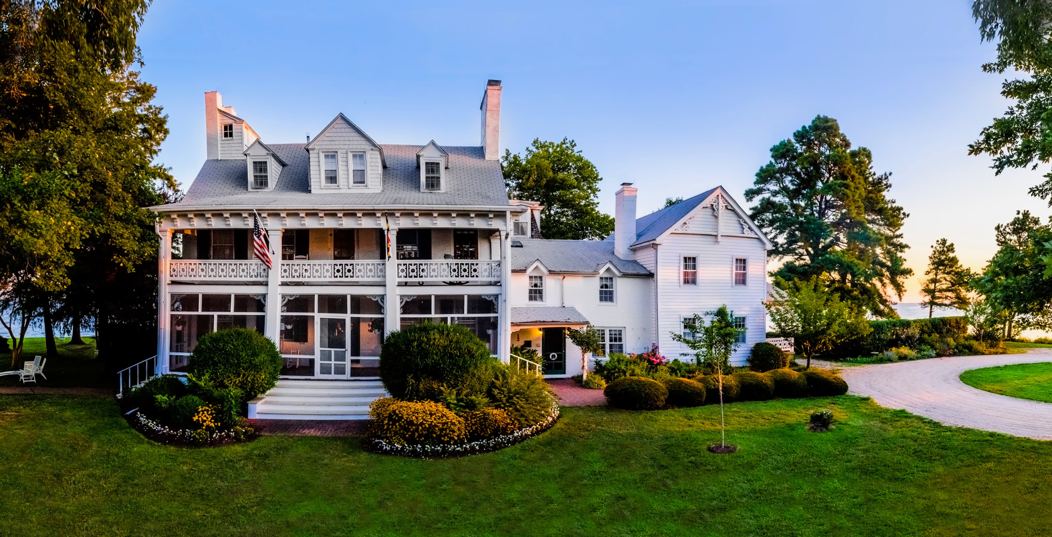 A large white house with a porch surrounded by lush greenery and landscaped gardens.