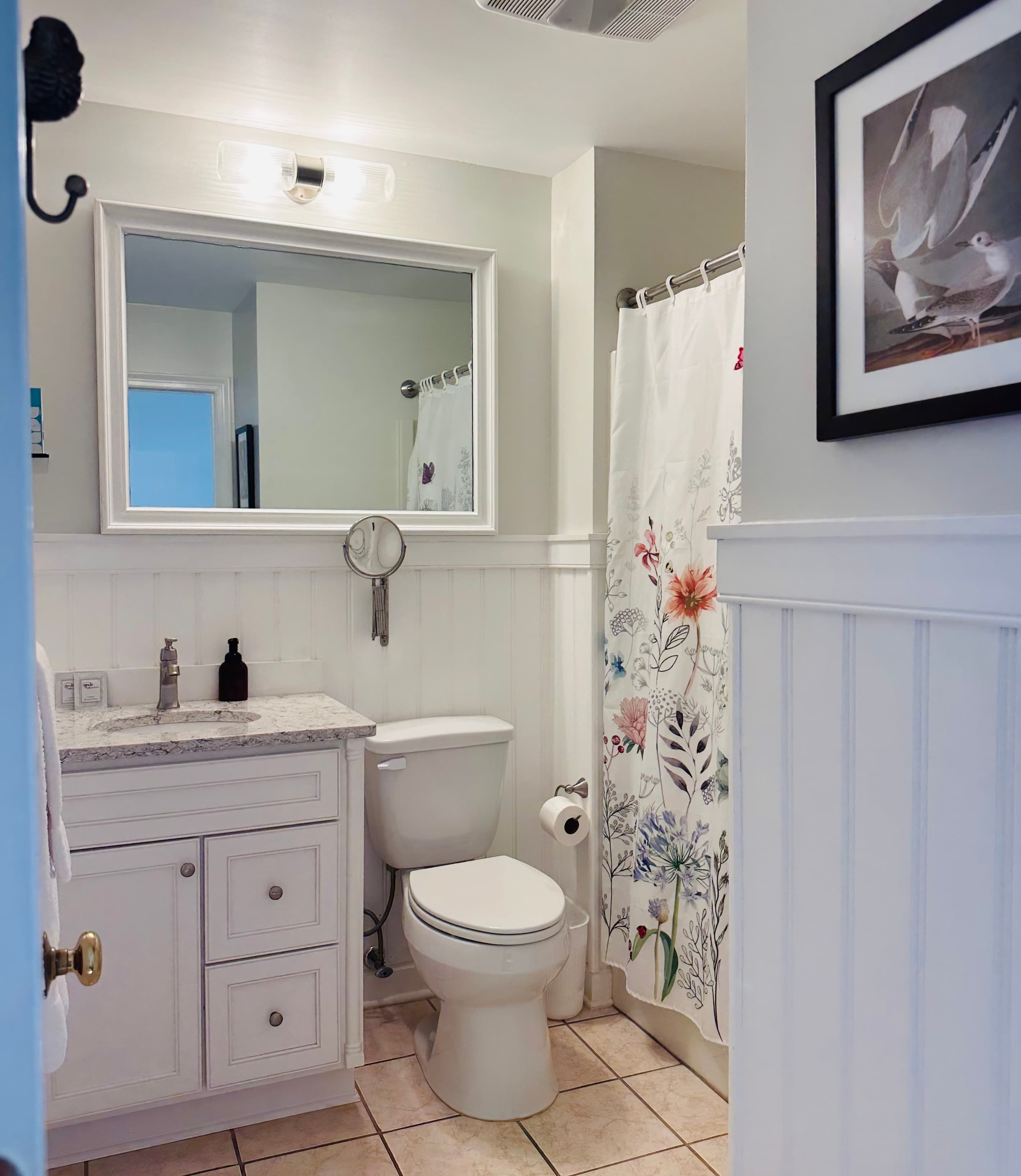 A small, bright bathroom with a floral shower curtain, white cabinetry, and a mirror reflecting the space.