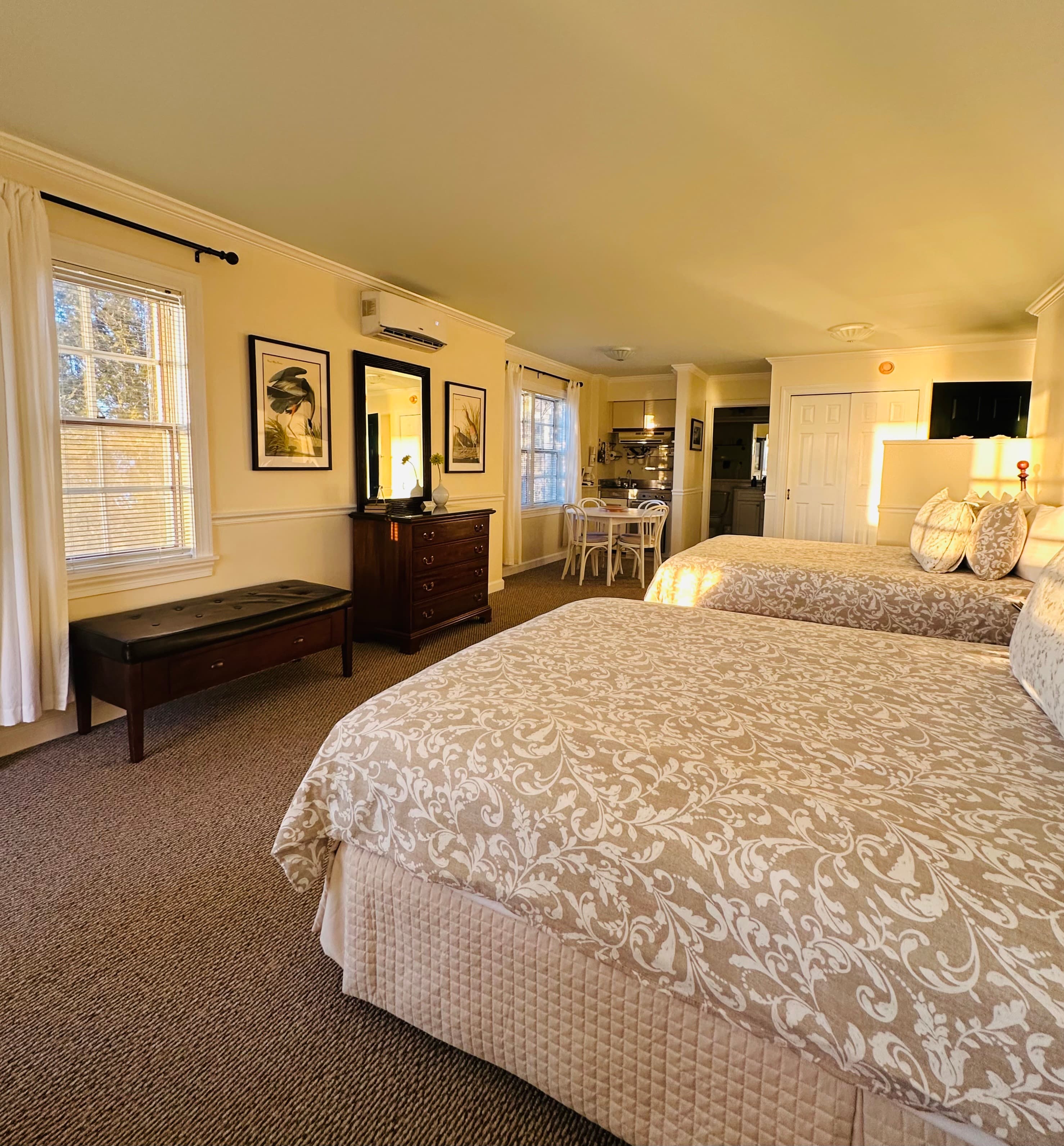A cozy hotel room featuring two beds, a dresser, a dining area, and natural light from windows.
