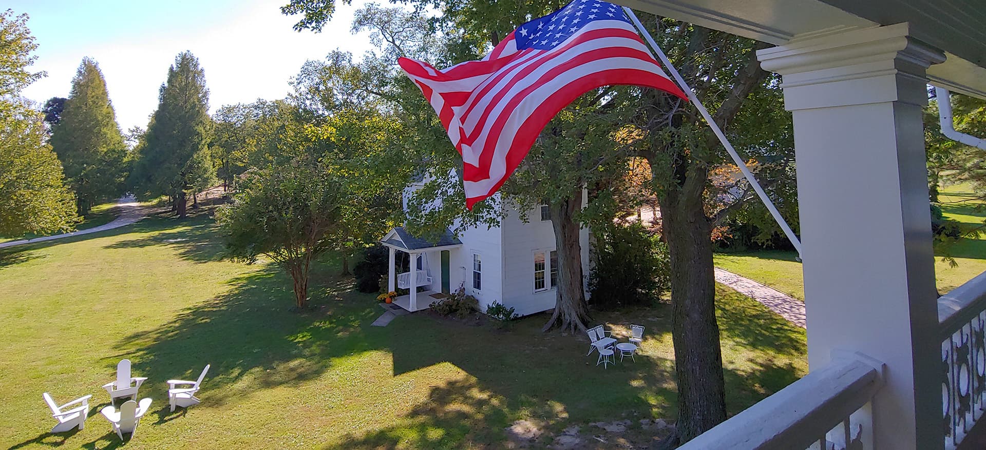 An American flag flutters in the breeze above a spacious green yard with a white house and outdoor seating.