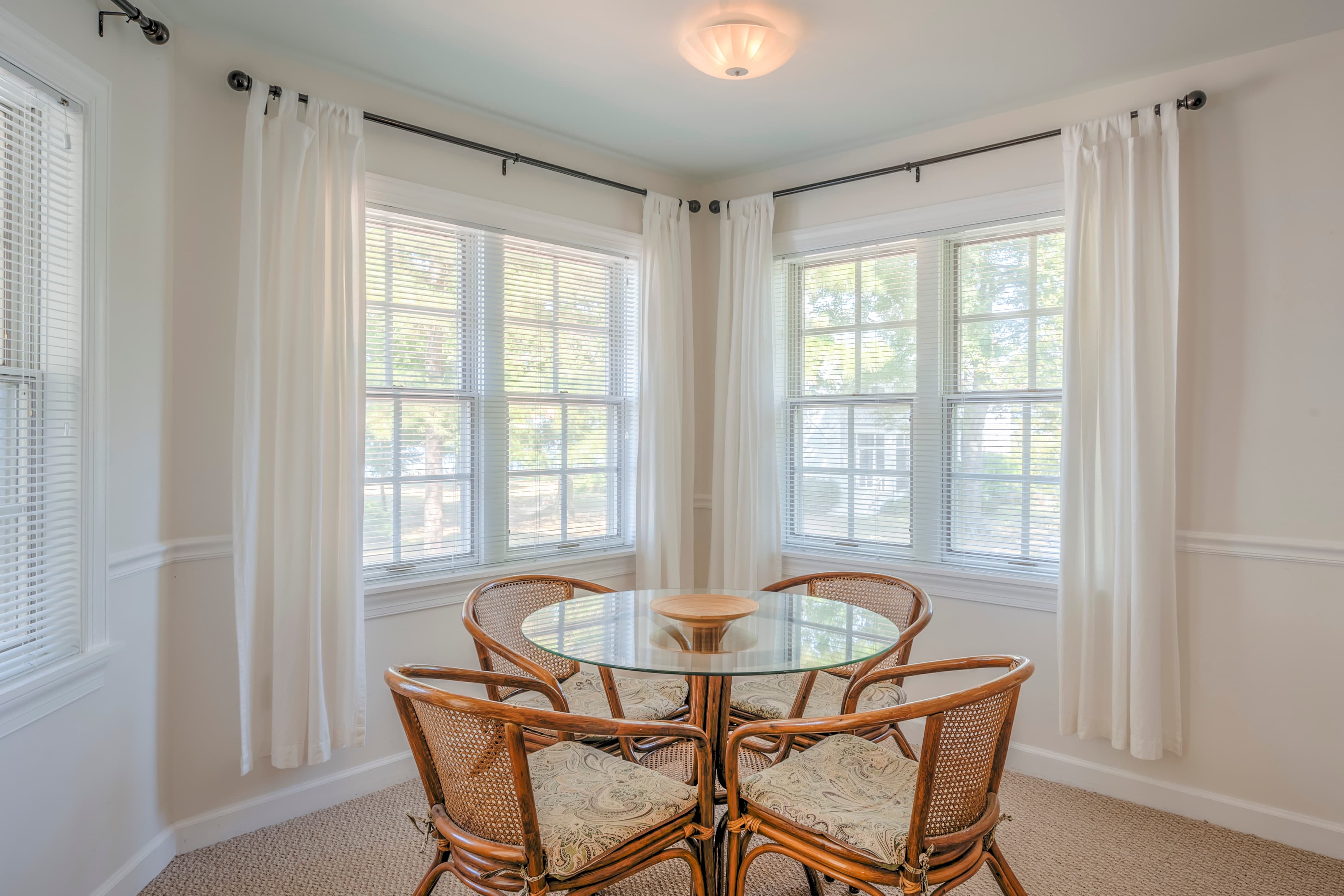 Bright corner dining area with a round glass table and four wicker chairs, surrounded by large windows.