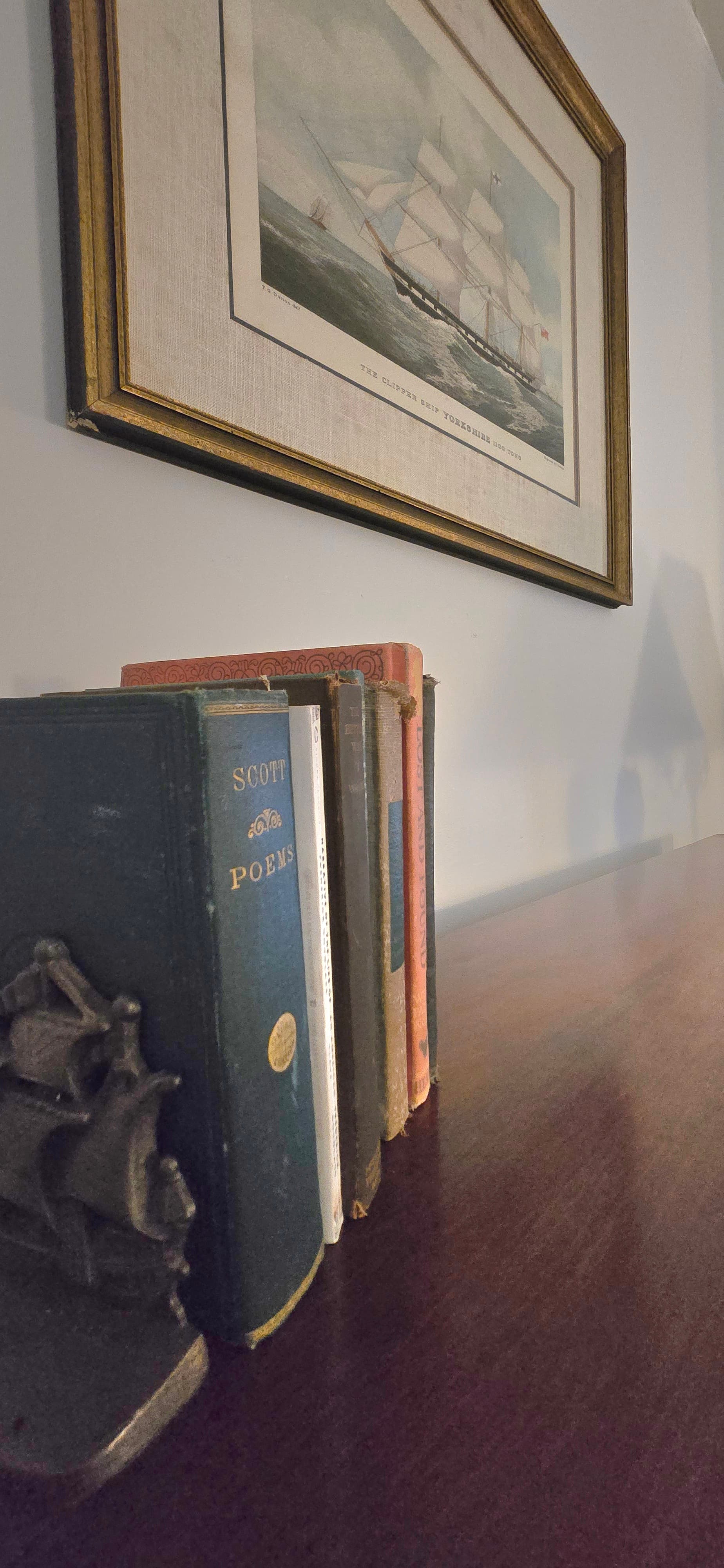 A decorative bookshelf displaying vintage books with a ship sculpture and a maritime painting on the wall.