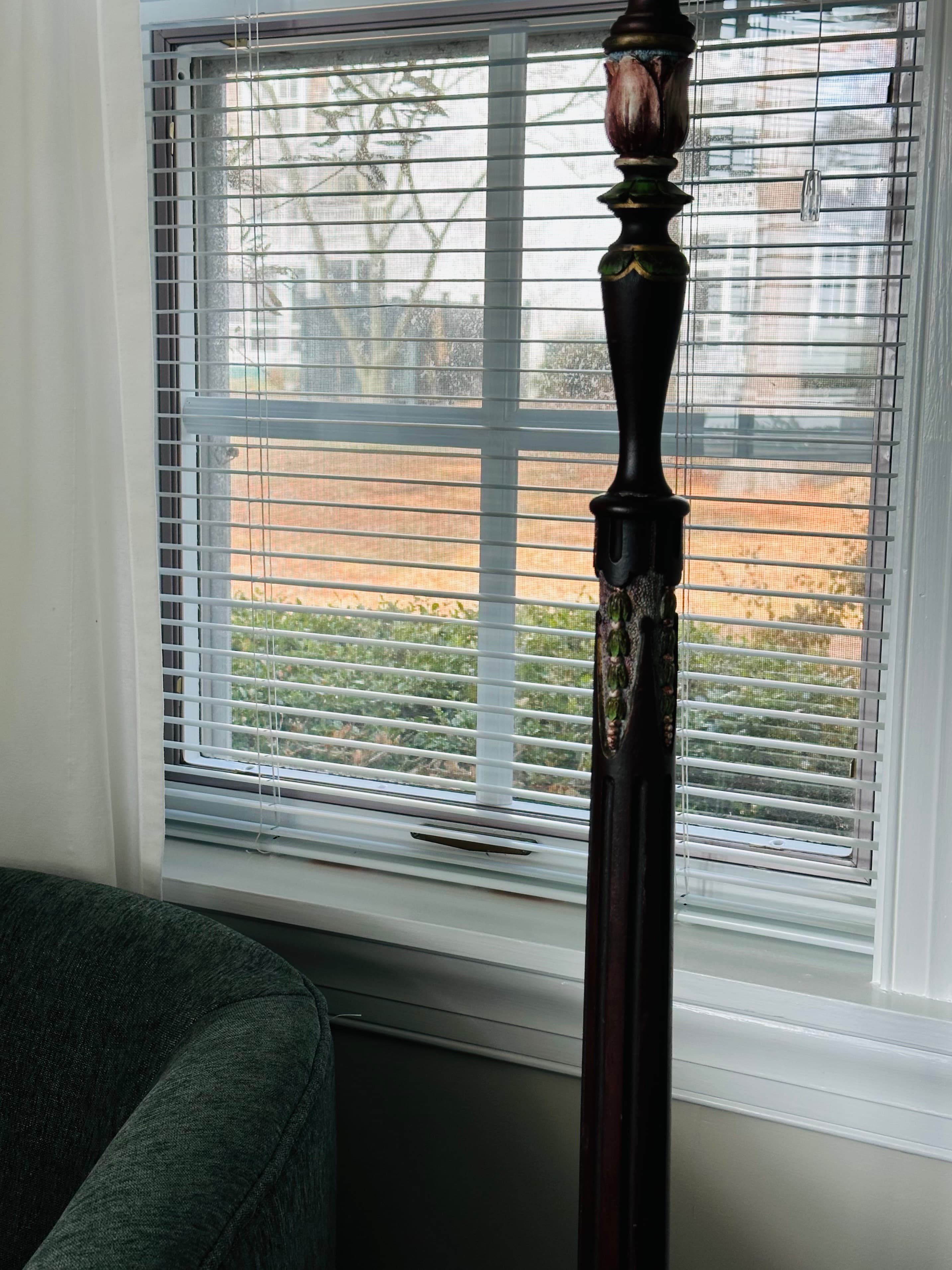 A wooden lamp stands next to a window with blinds, casting light onto a nearby green sofa.