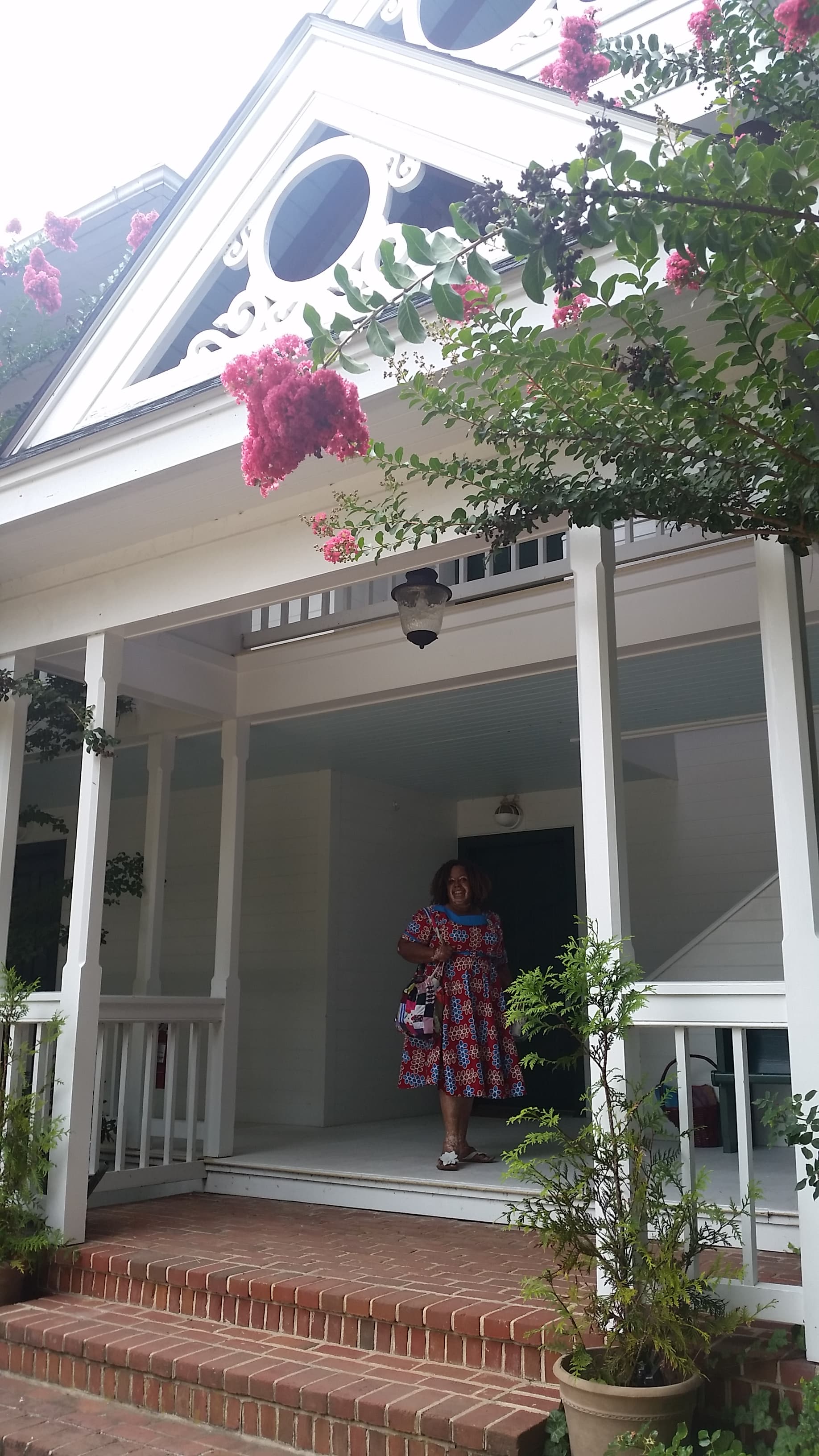 A woman in a colorful dress stands on the porch of a house adorned with pink flowers.
