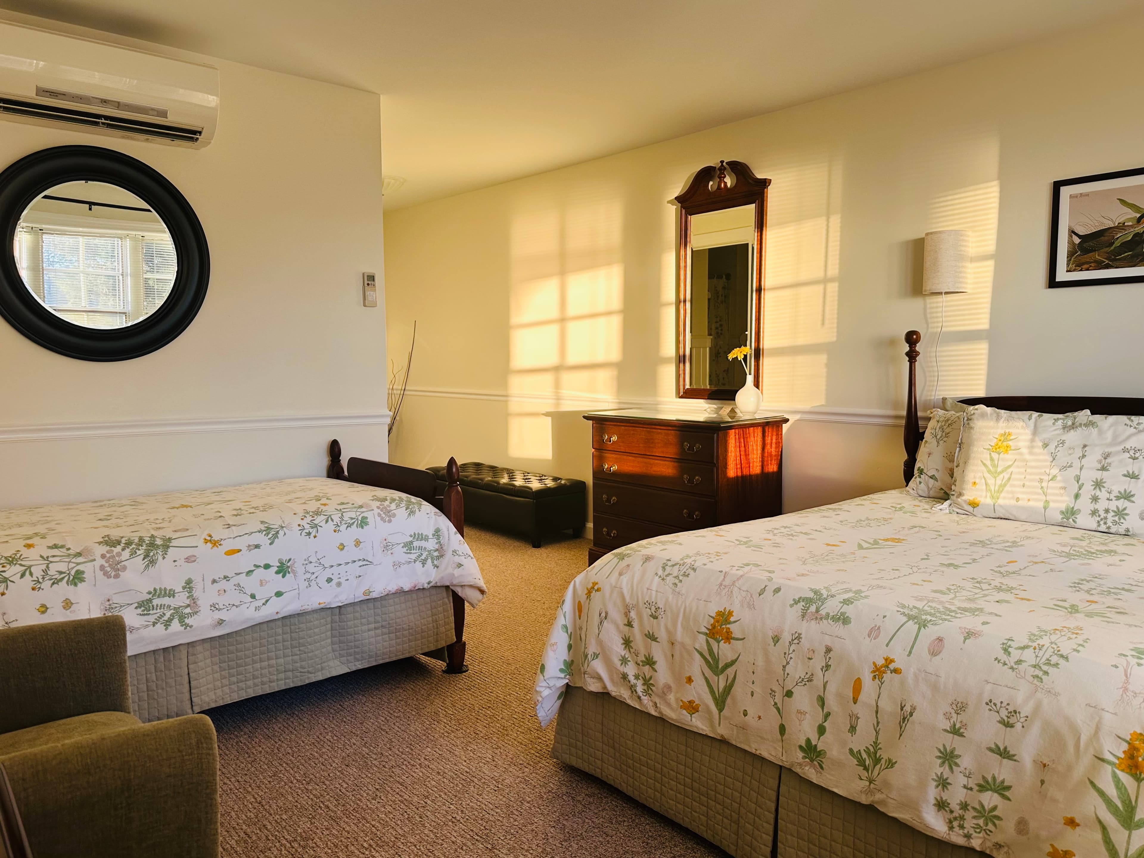 A sunlit bedroom featuring two floral-patterned beds, a dresser, and a round mirror.
