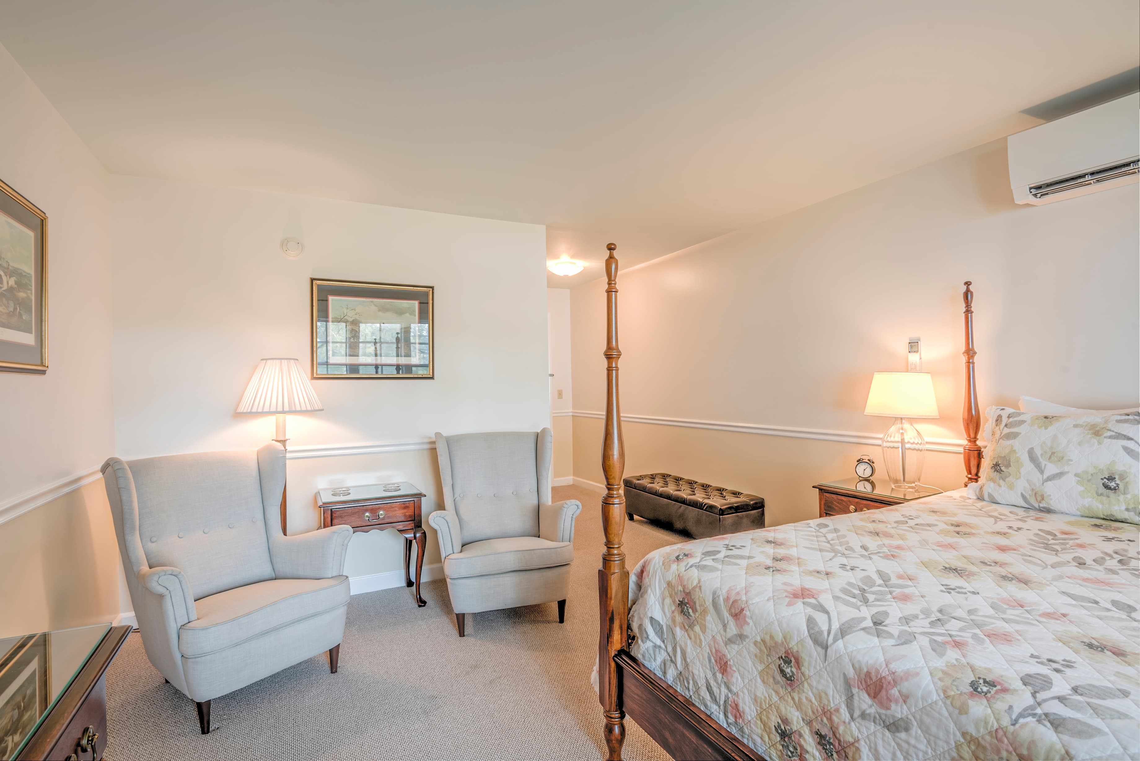 A cozy bedroom featuring a floral quilted bedspread, two armchairs, and warm lighting.