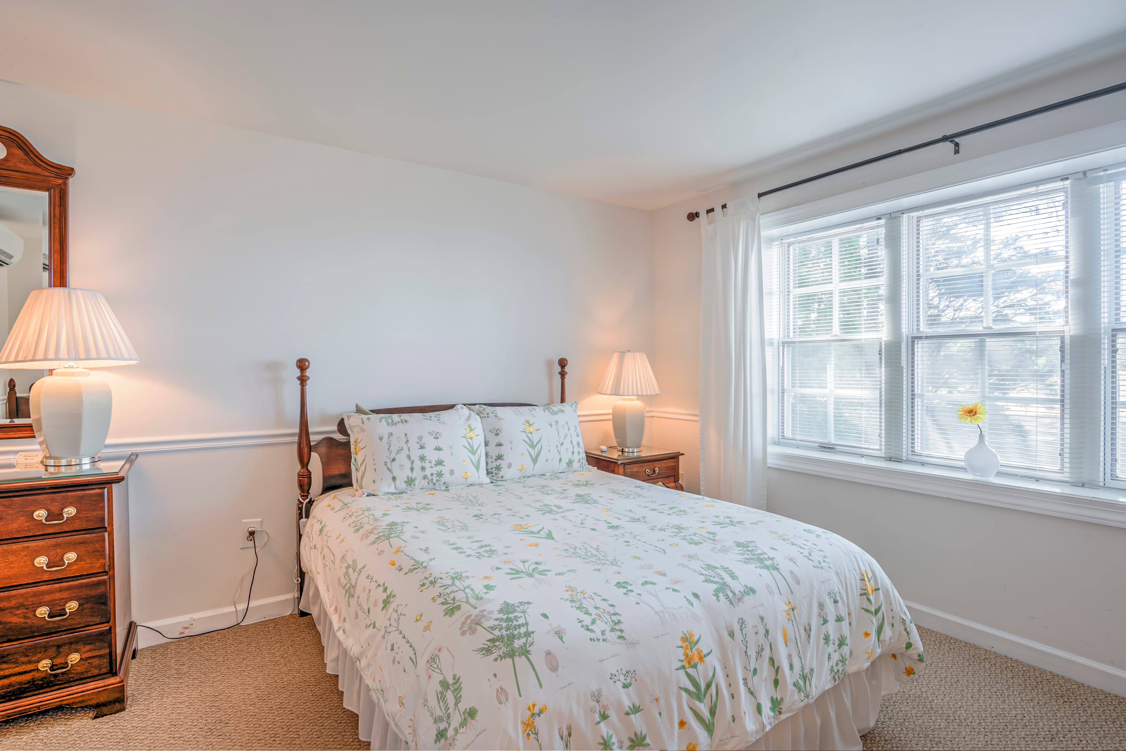 Cozy bedroom featuring a floral bedspread, wooden furniture, and natural light from a window.