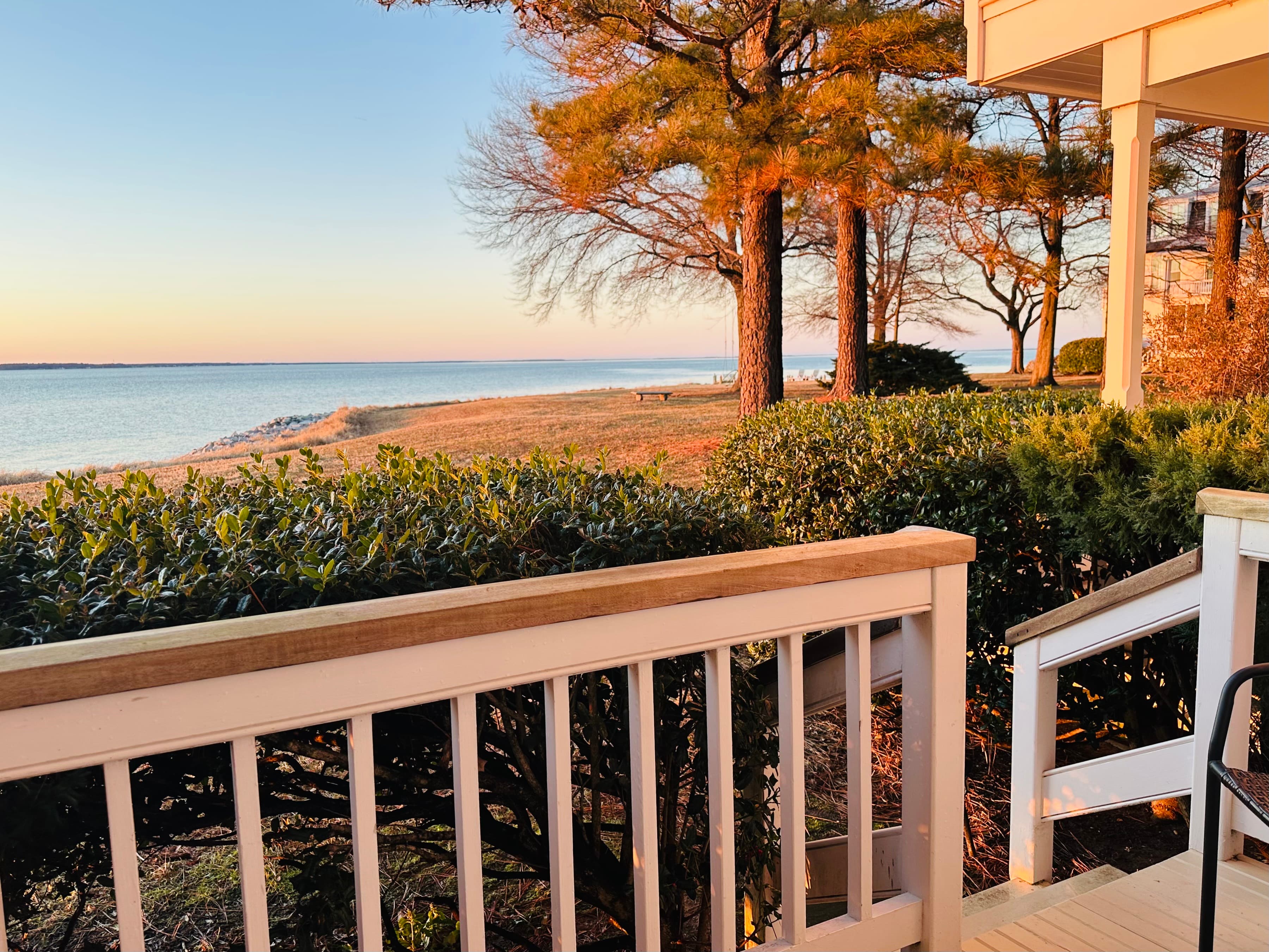 A tranquil view of a coastal landscape with trees and a soft sunset glow from a porch railing.