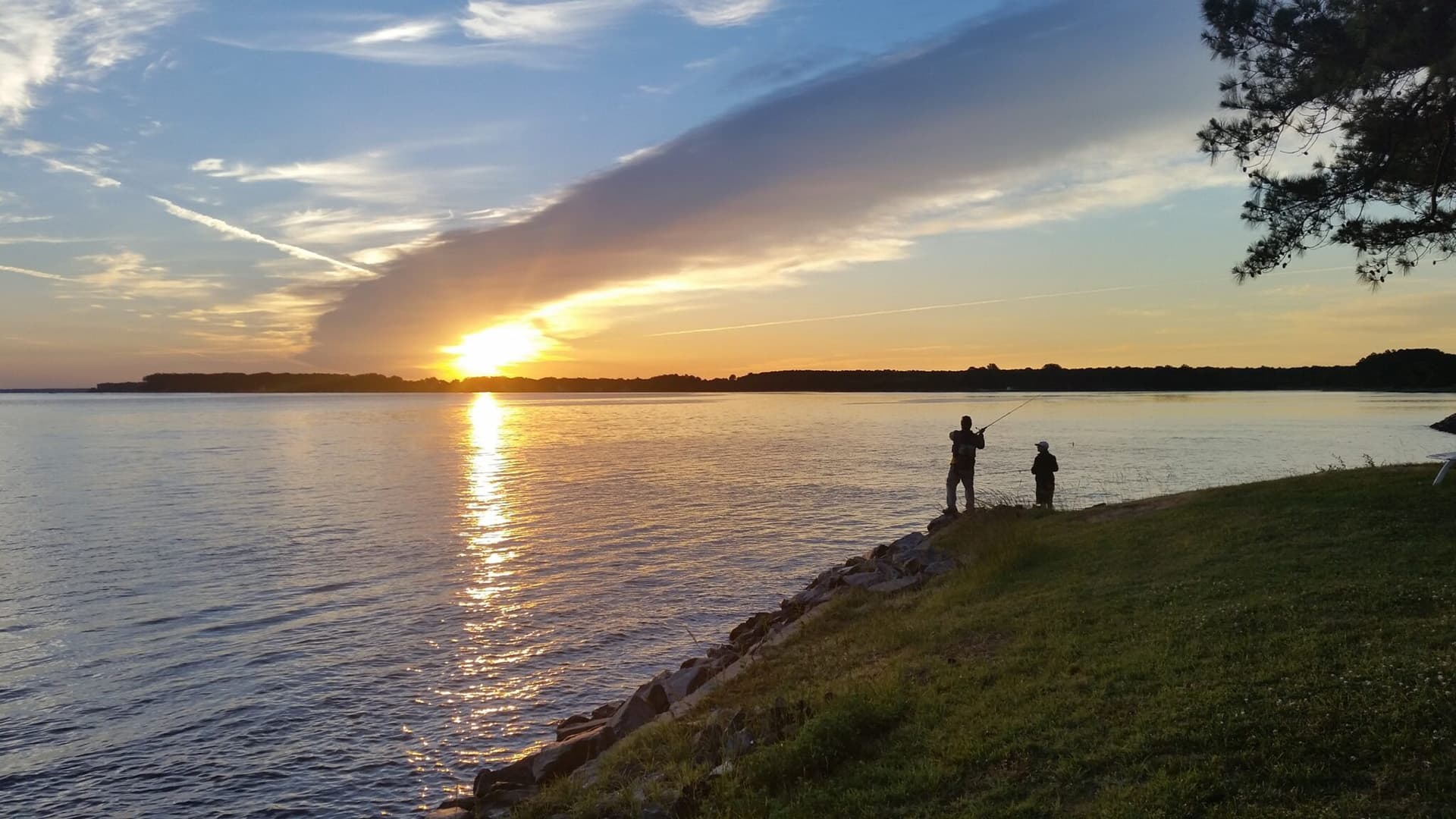 Two people stand by a lake at sunset, casting shadows against the shimmering water.