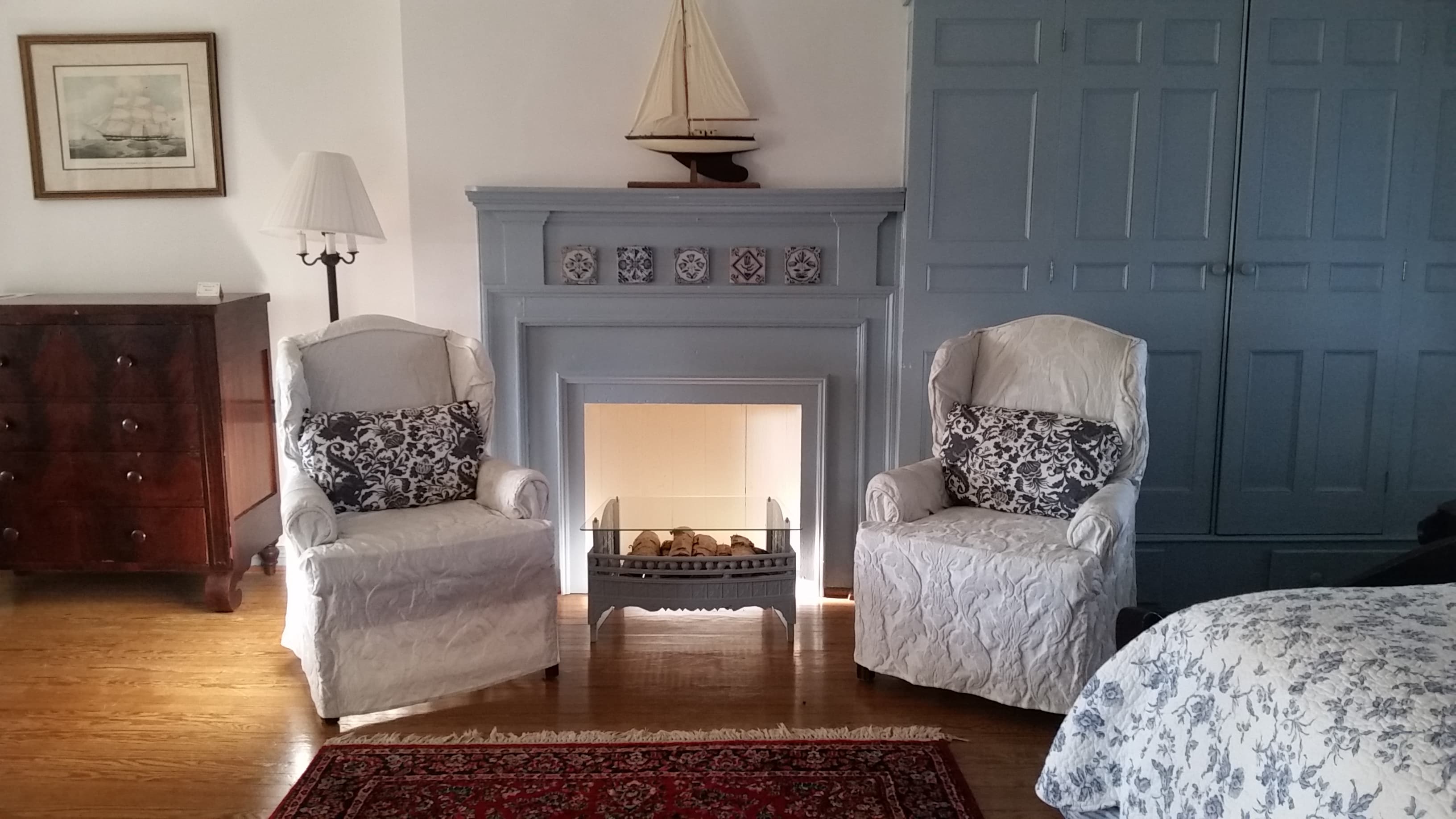 A cozy room featuring two white upholstered chairs, a simple fireplace, and a wooden dresser.