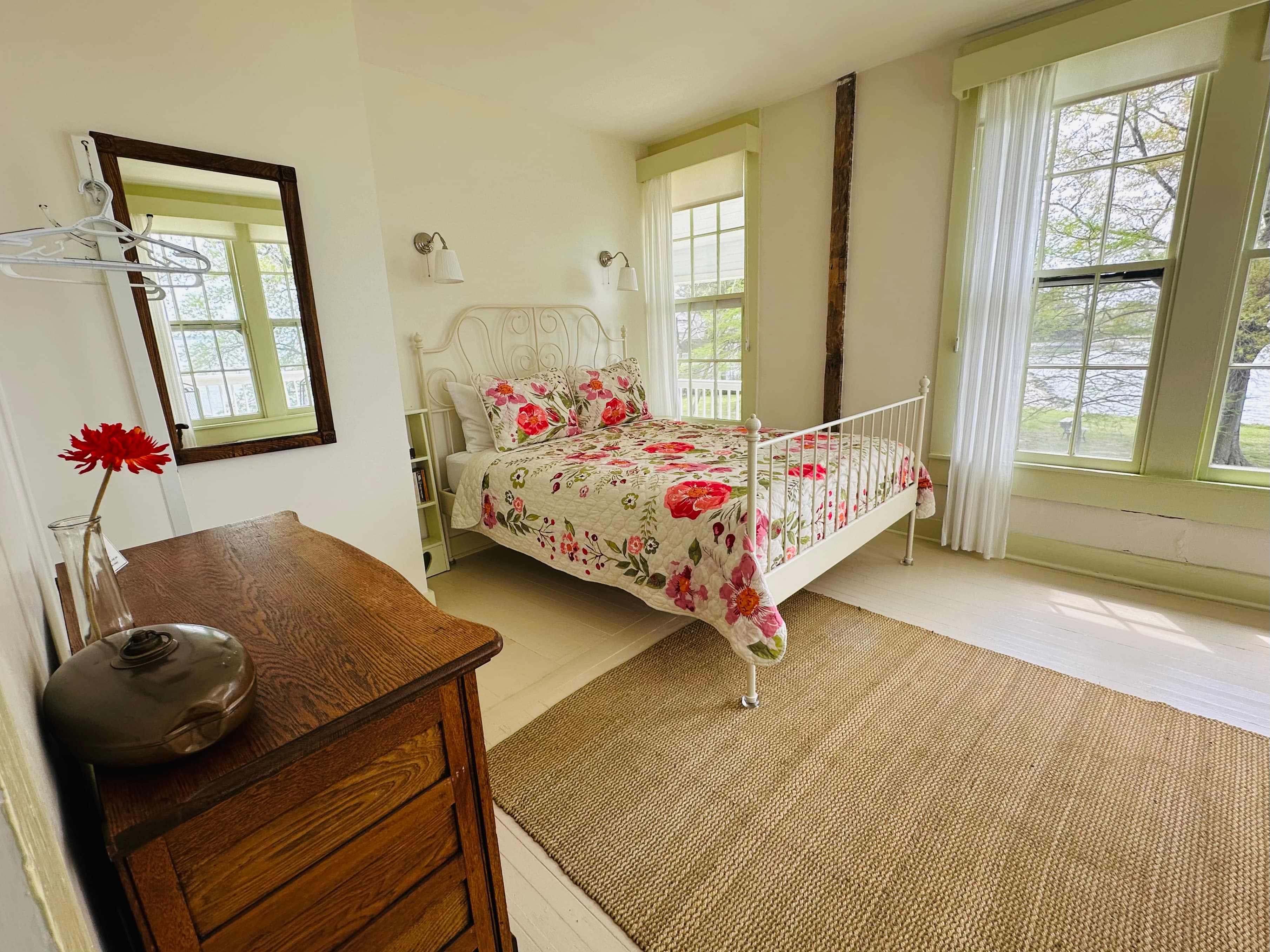 A cozy bedroom featuring a white metal bed with floral bedding, a wooden dresser, and large windows with views.
