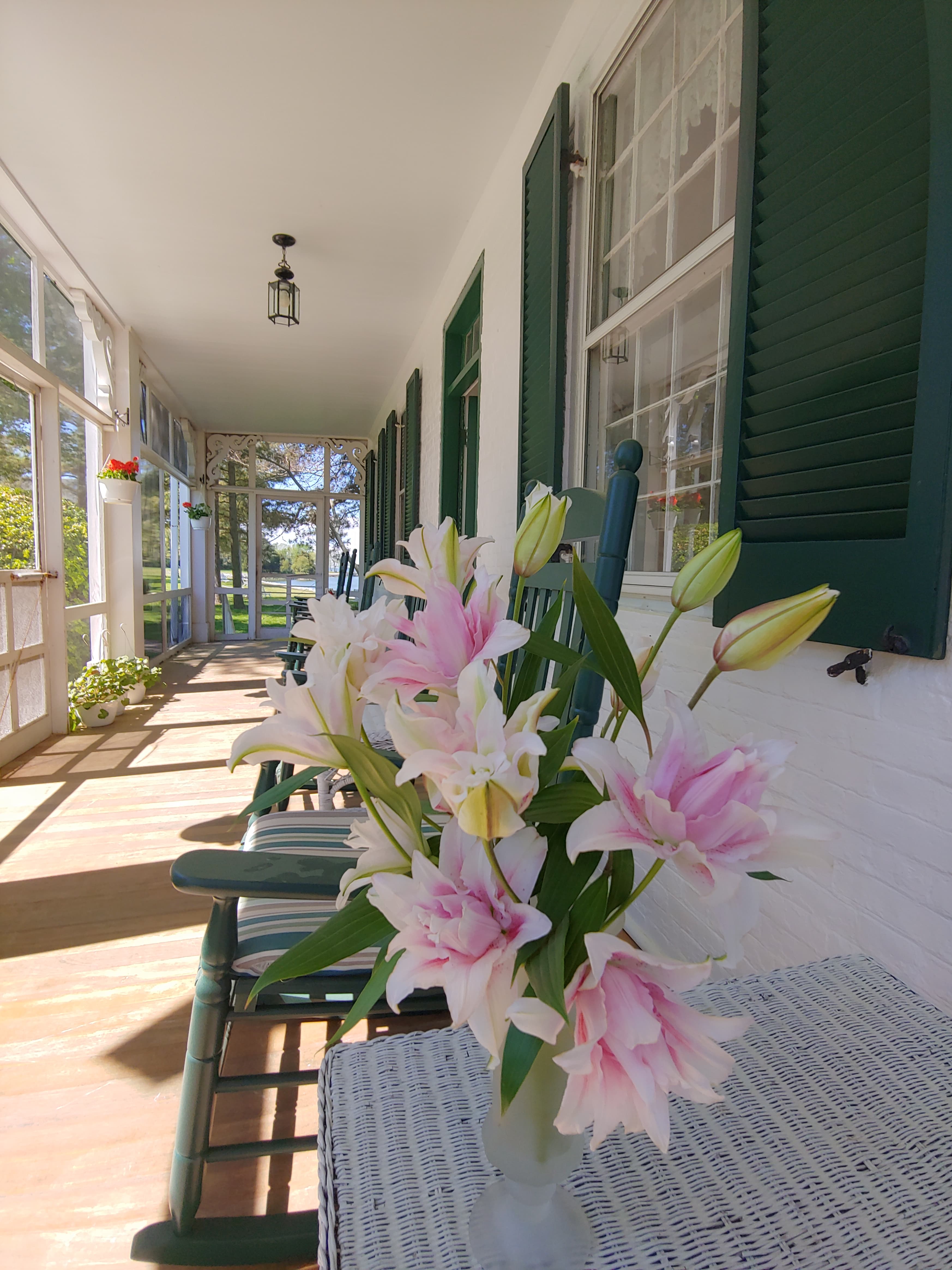 A bouquet of pink and white lilies sits on a table on a sunny porch with rocking chairs and green shutters.