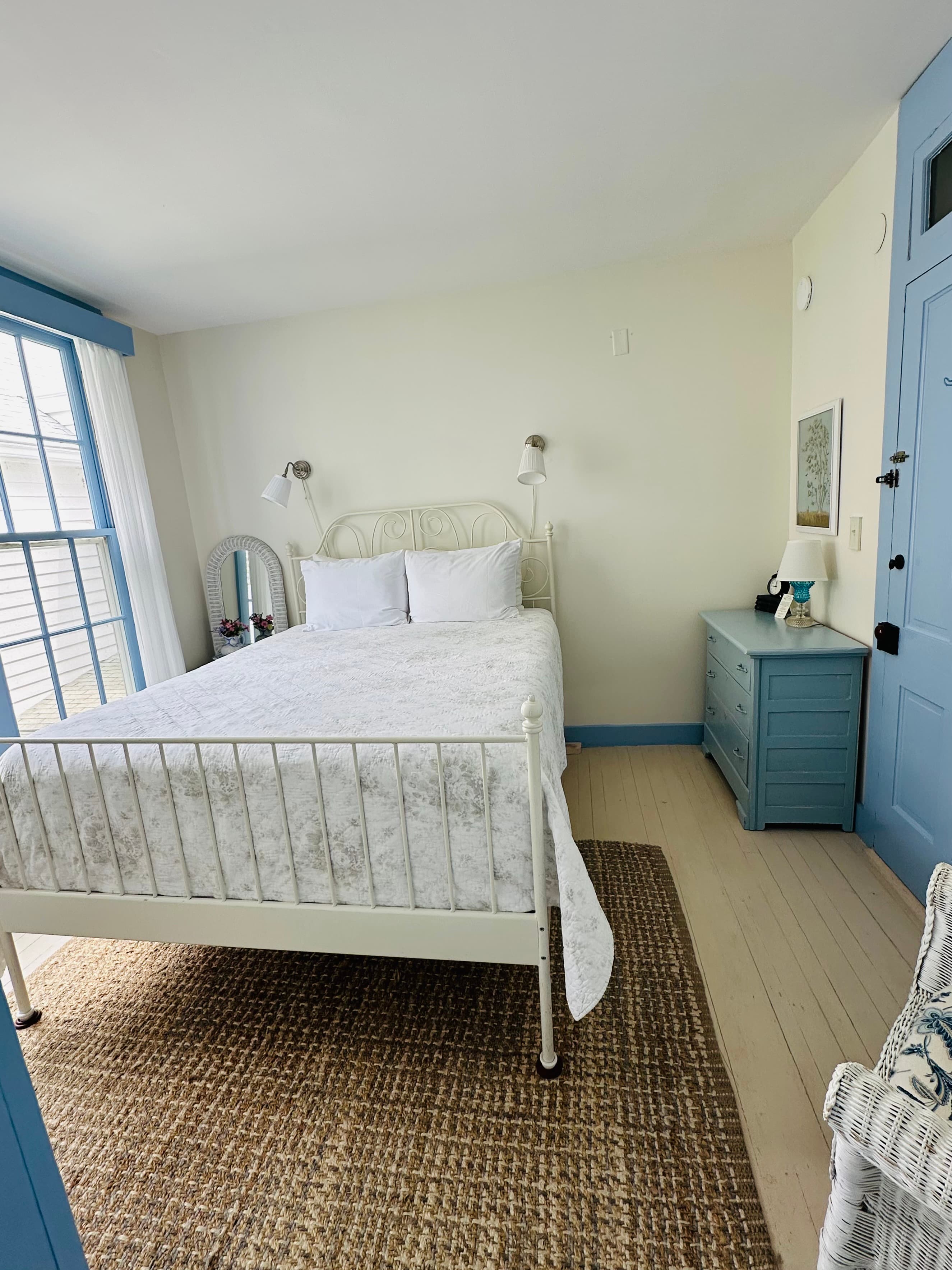Bright and airy bedroom featuring a white metal bed, light-colored walls, blue accents, and natural light from a window.