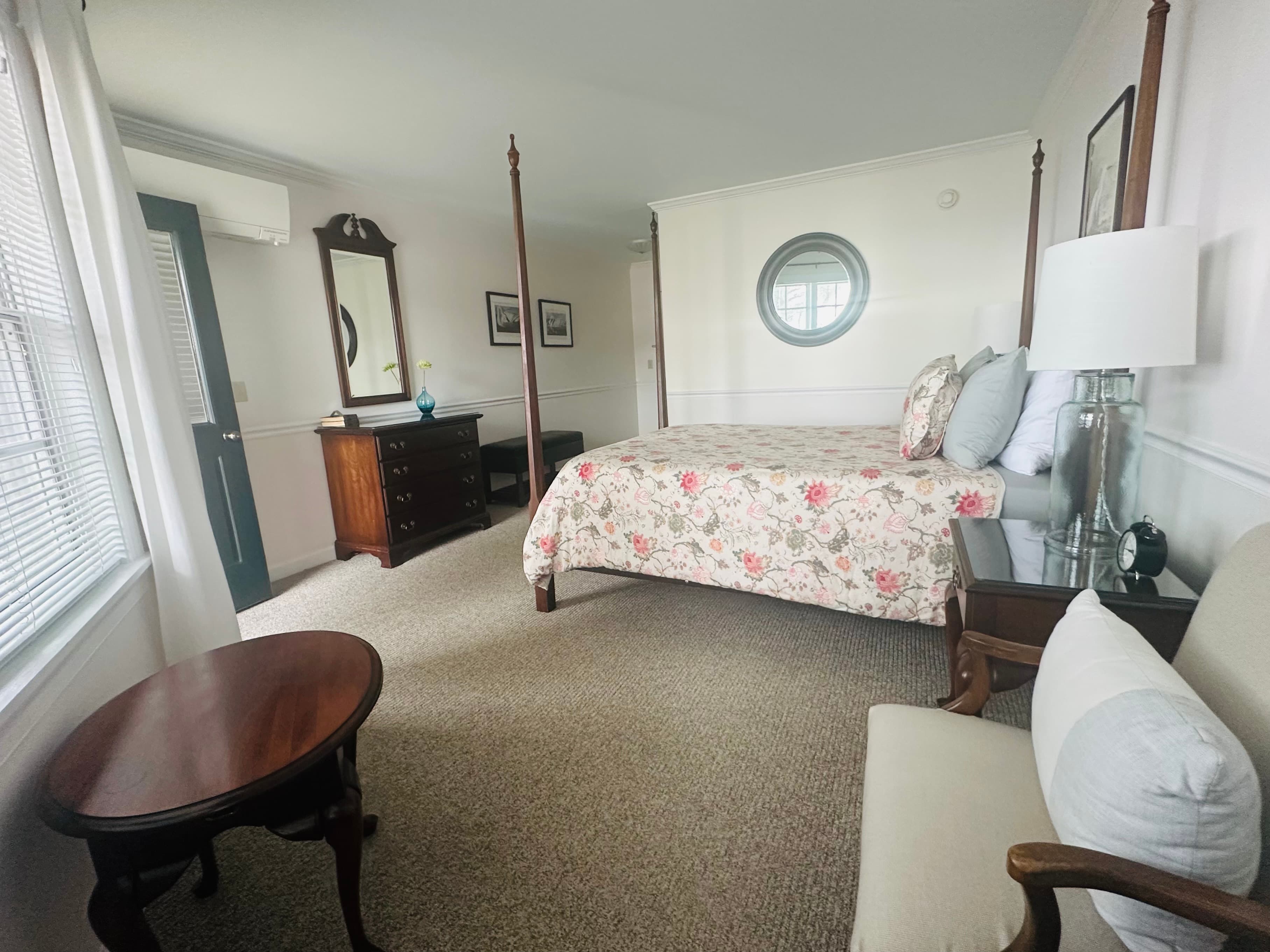 A cozy bedroom featuring a floral-patterned bedspread, vintage furniture, and natural light from a window.