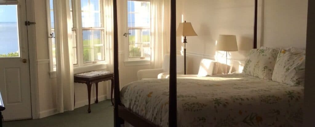 A bright, airy bedroom featuring a four-poster bed, side table, and large windows with a view.