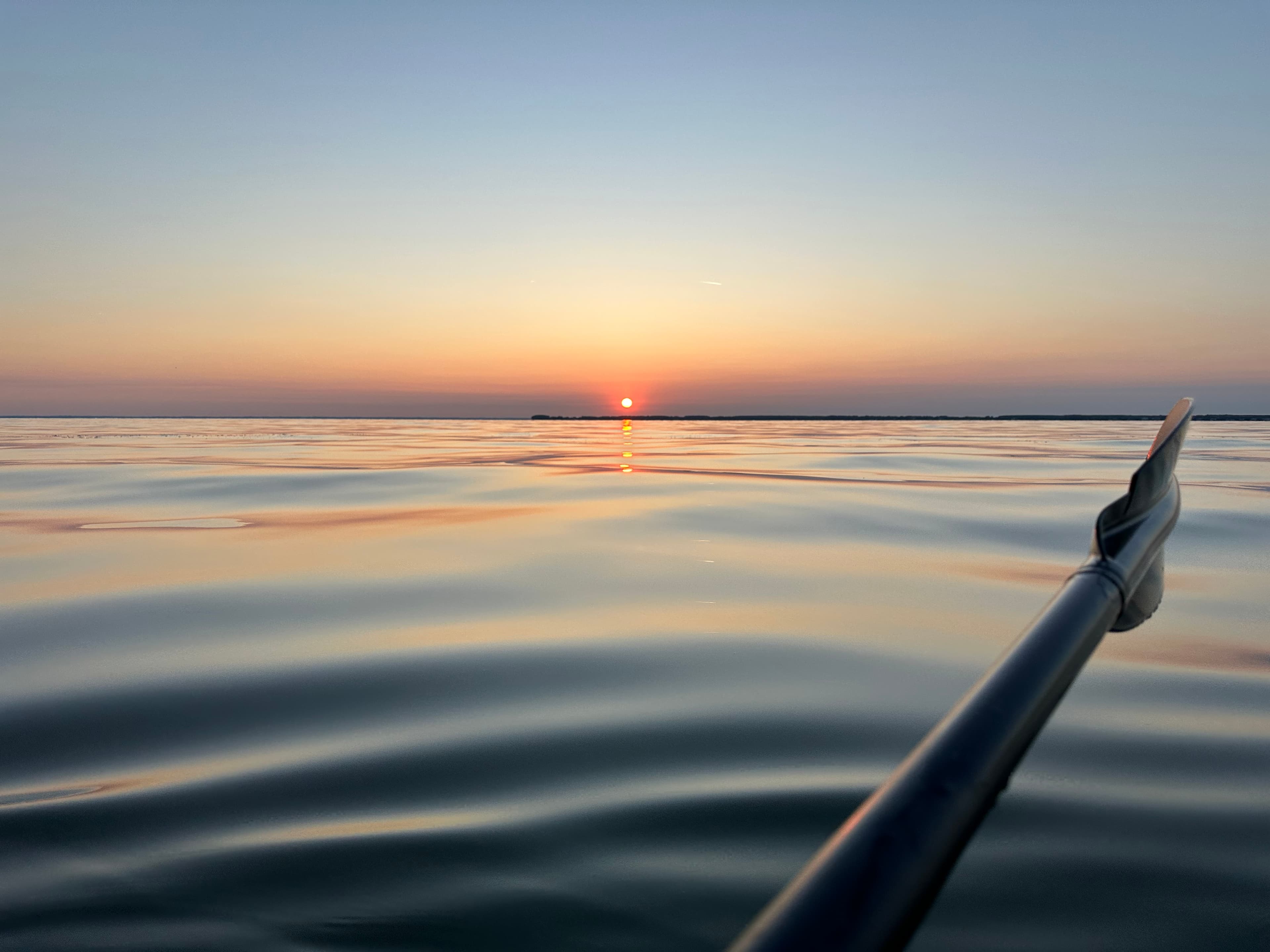 A paddle extends into calm water as the sun sets on the horizon, casting a warm glow over the scene.