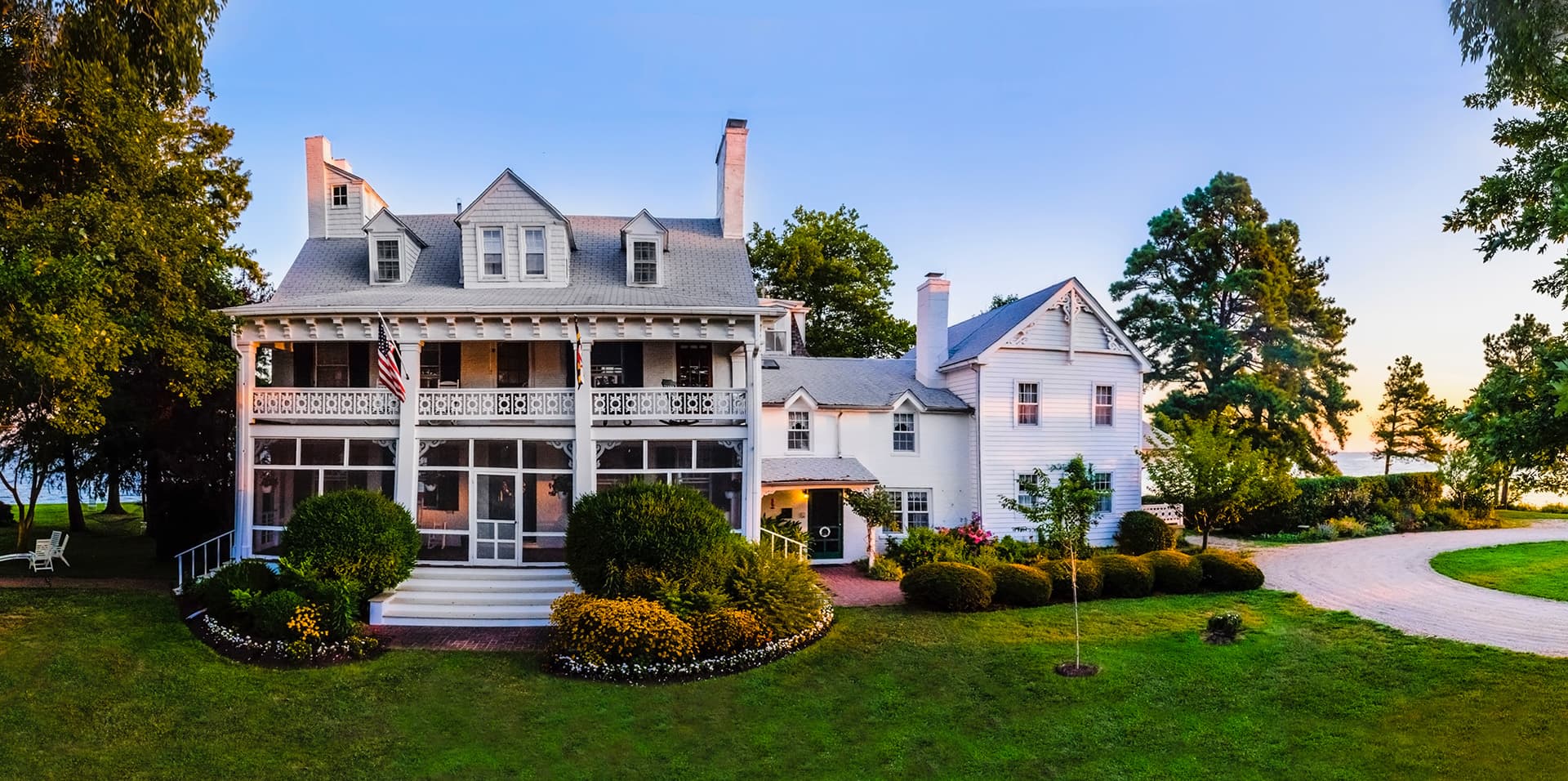 A large, white, historic house with a porch and manicured lawn surrounded by trees and flowers.