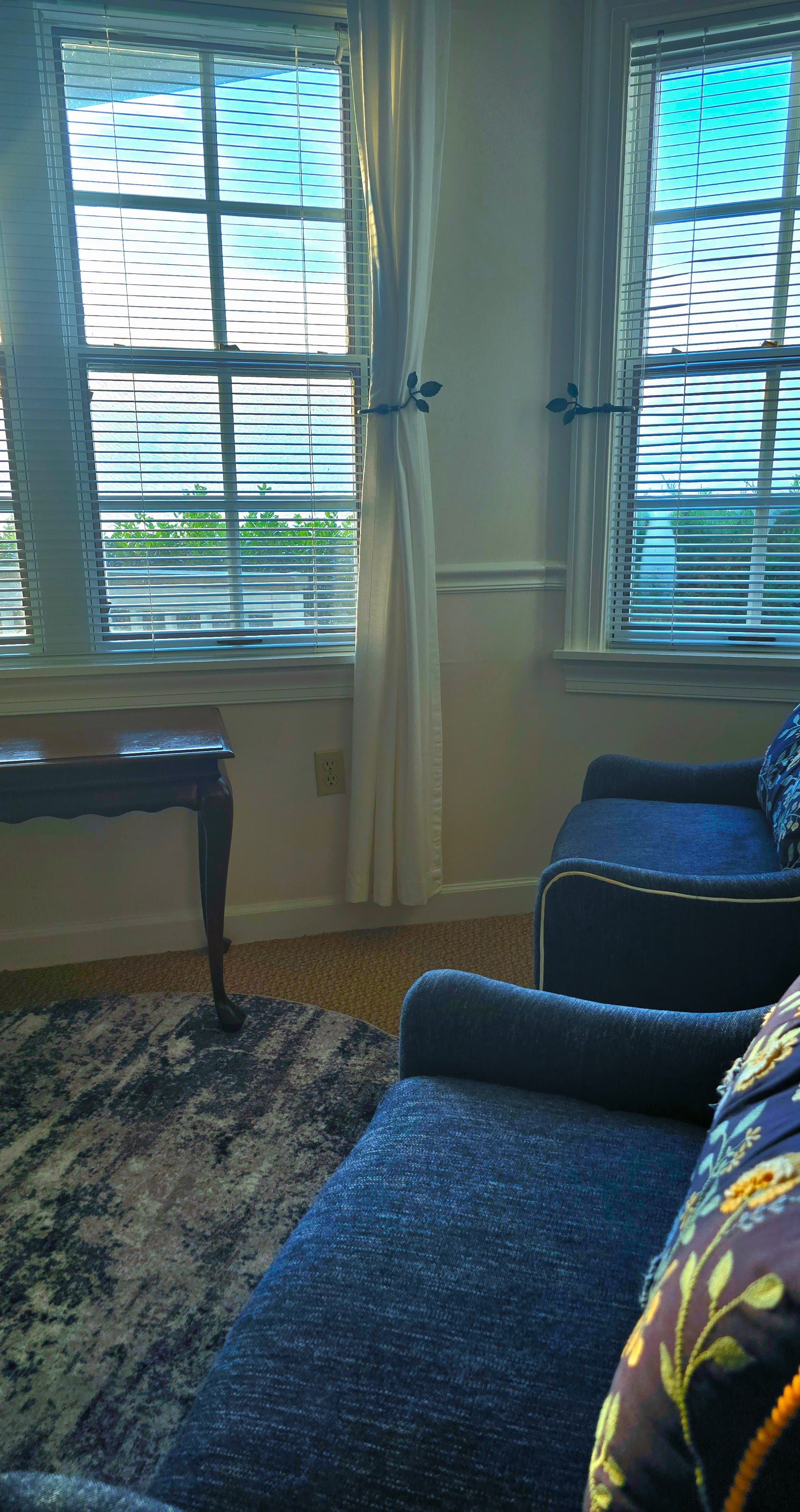A cozy sitting area with blue chairs, a small table, and windows draped with light curtains.