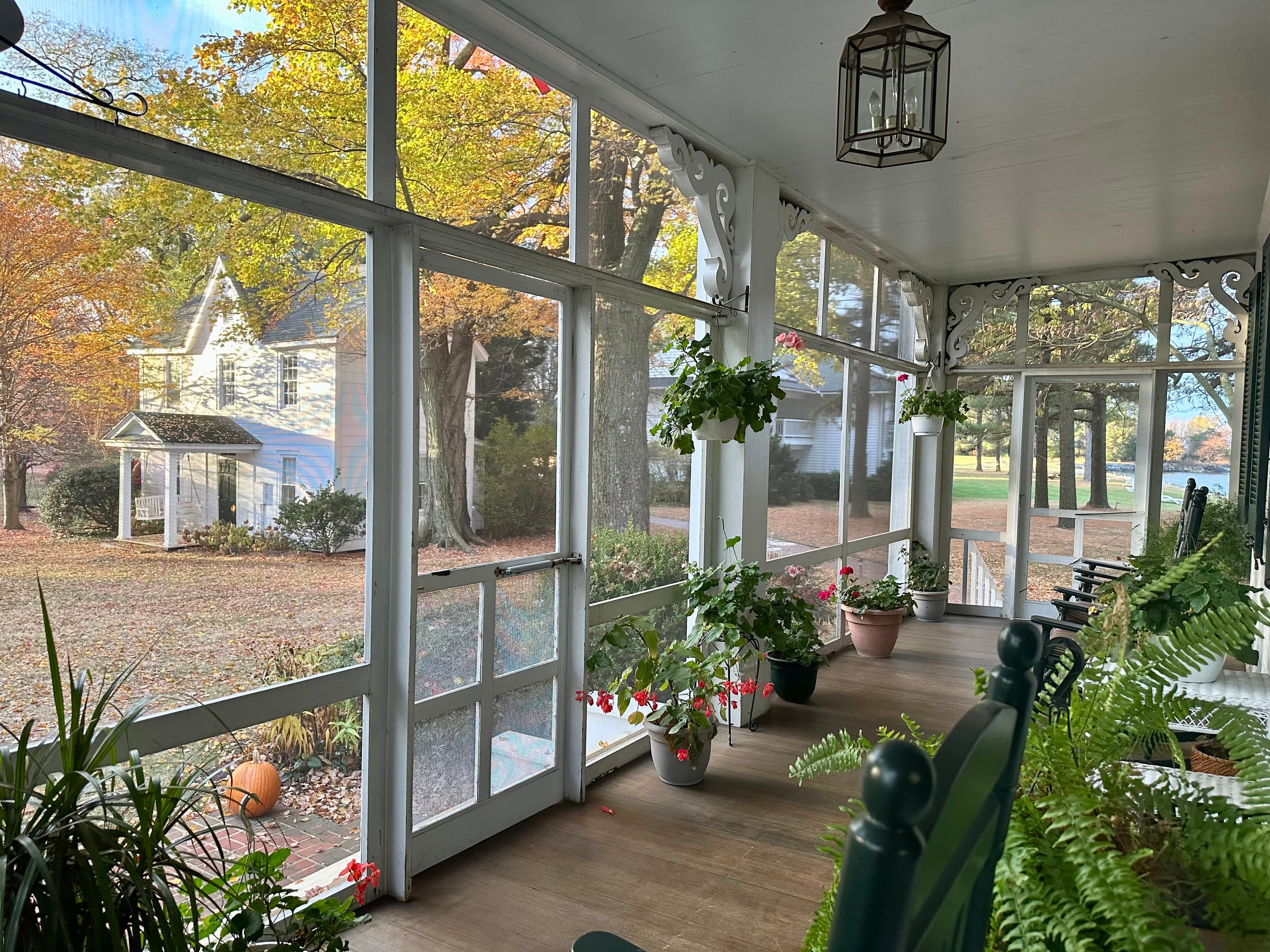 A sunlit porch with hanging plants overlooks a colorful autumn landscape and a white house in the background.
