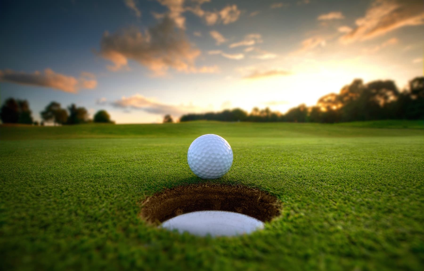 A golf ball is poised near a hole on a lush green course at sunset. A golf ball is poised near a hole on a lush green course at sunset.