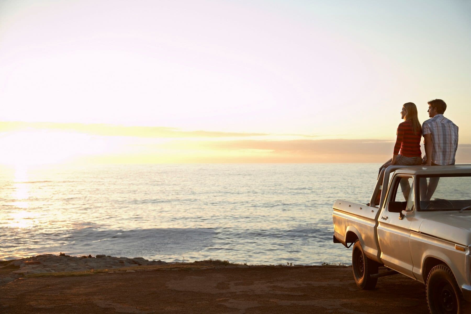 Couple sitting on the bed of a truck looking out at a sunset over the water.